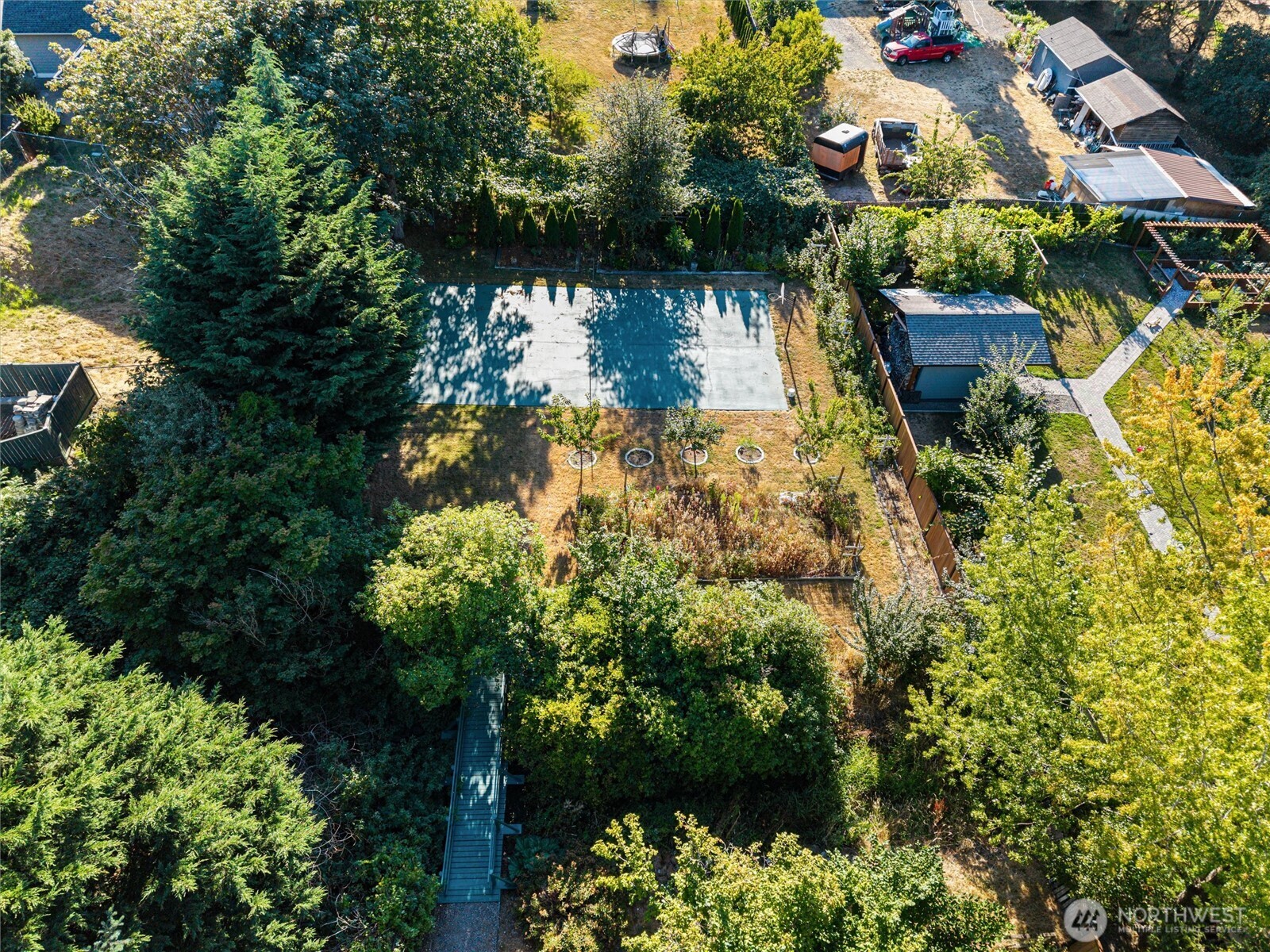 Aerial view of a wooded backyard with a large concrete pad, garden beds, and a footbridge.