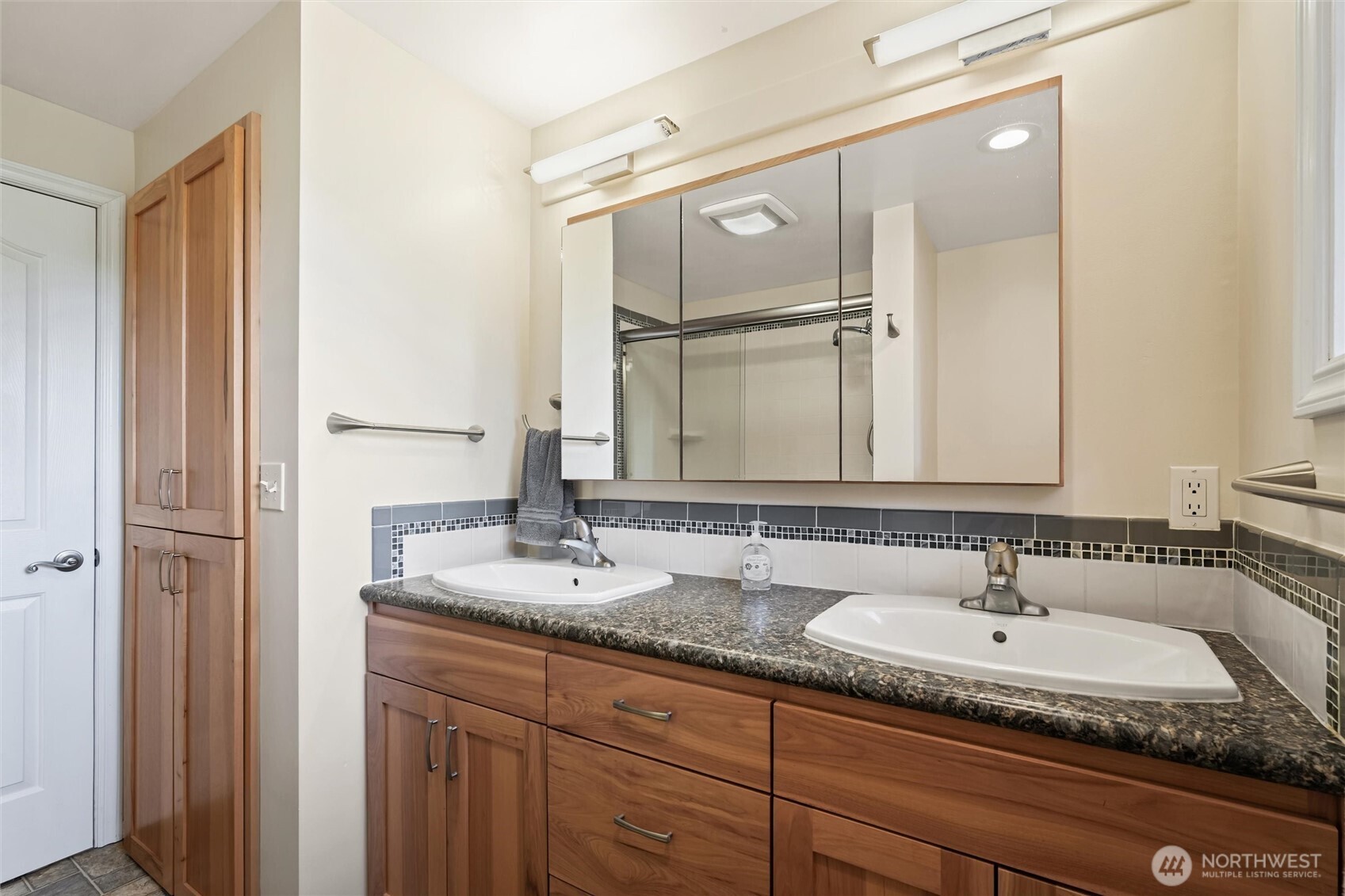 Bathroom double vanity with warm wood cabinets, dark stone countertop, and large mirrored medicine cabinet.
