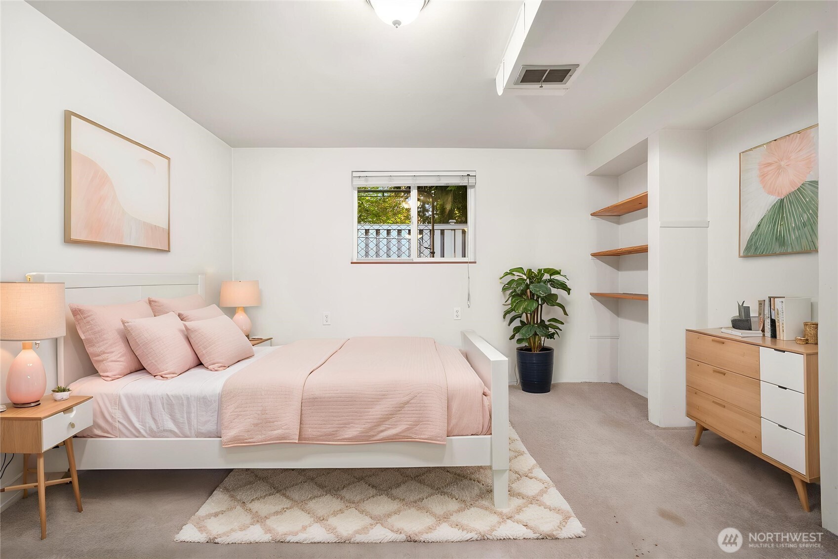 Staged bedroom with a white bed in pale pink bedding, built-in wood shelves, and a window overlooking greenery.