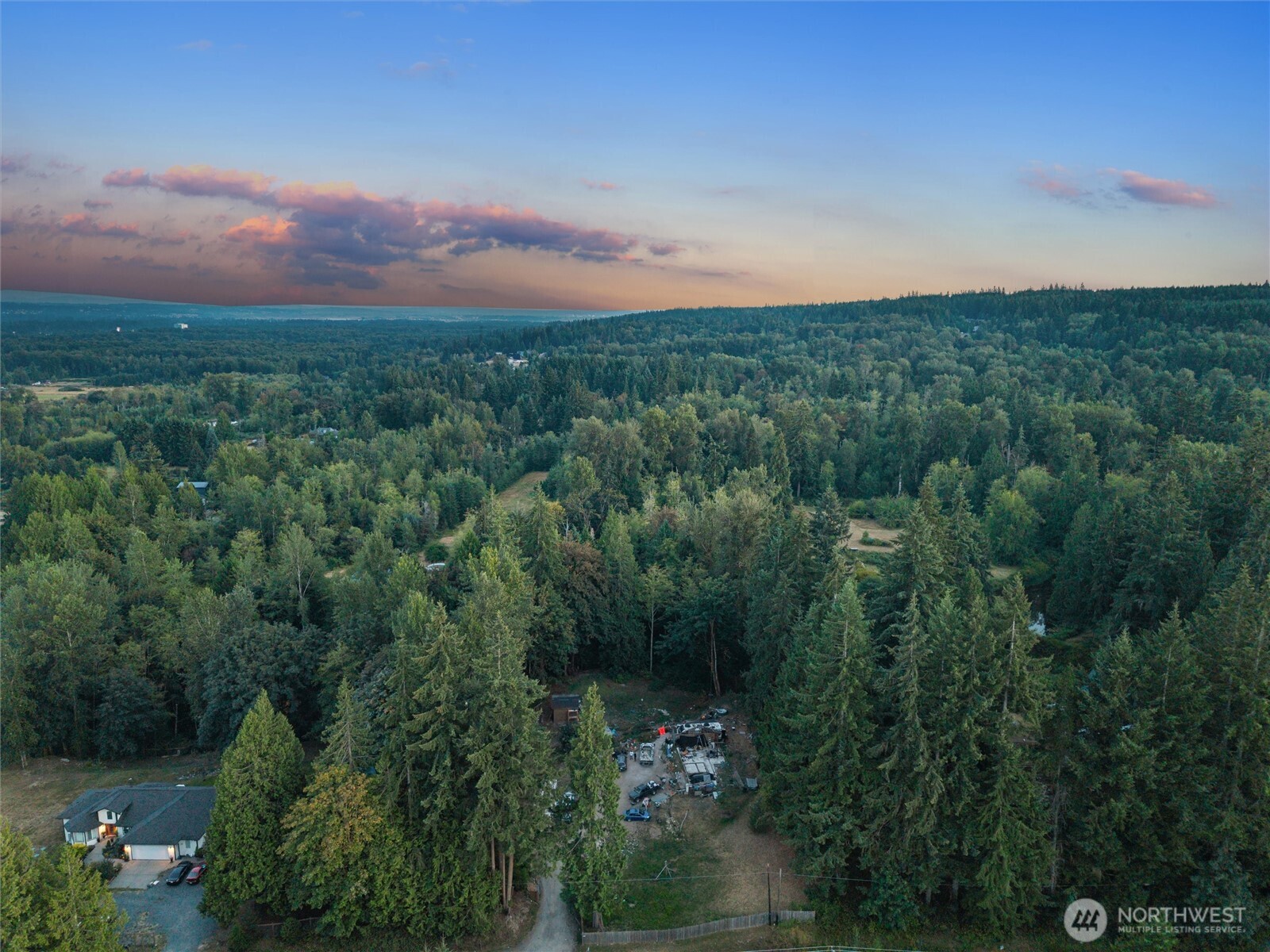 Aerial view of a wooded property with a clearing, neighboring home, and distant water at dusk.