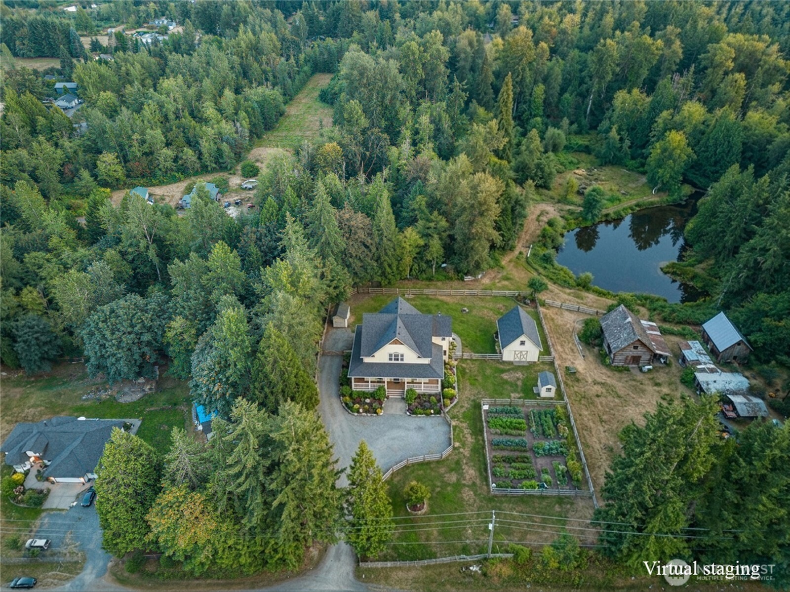 Aerial view of a wooded property with a house, outbuildings, fenced gardens, and a pond.