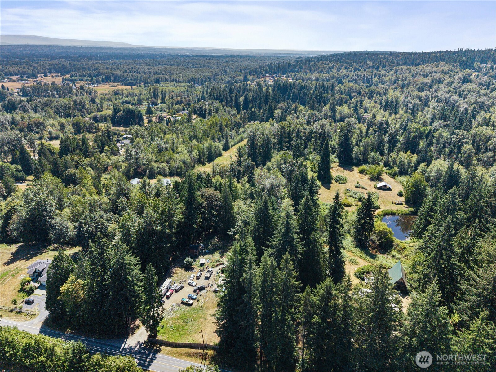 Aerial view of a wooded property with scattered outbuildings, clearings, a pond, and a road.