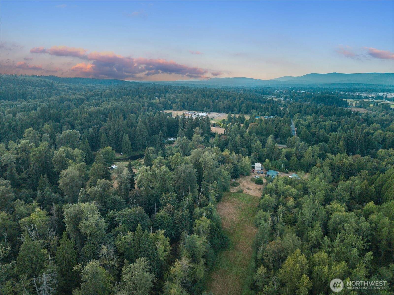 Aerial view of a wooded property with a grassy clearing and distant ridges beneath pink clouds.