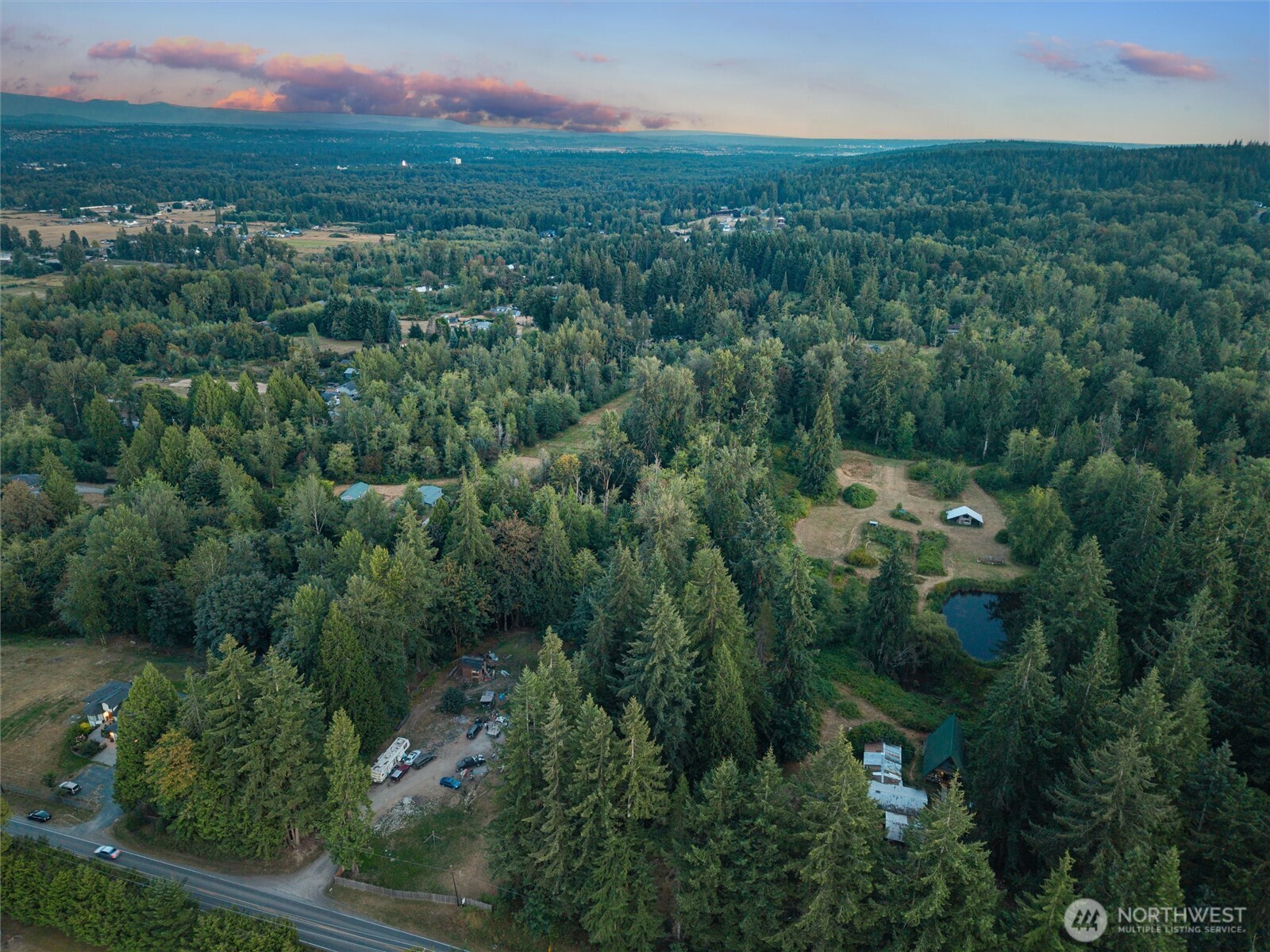Aerial view of wooded property with scattered buildings, a small pond, and a road in the foreground.