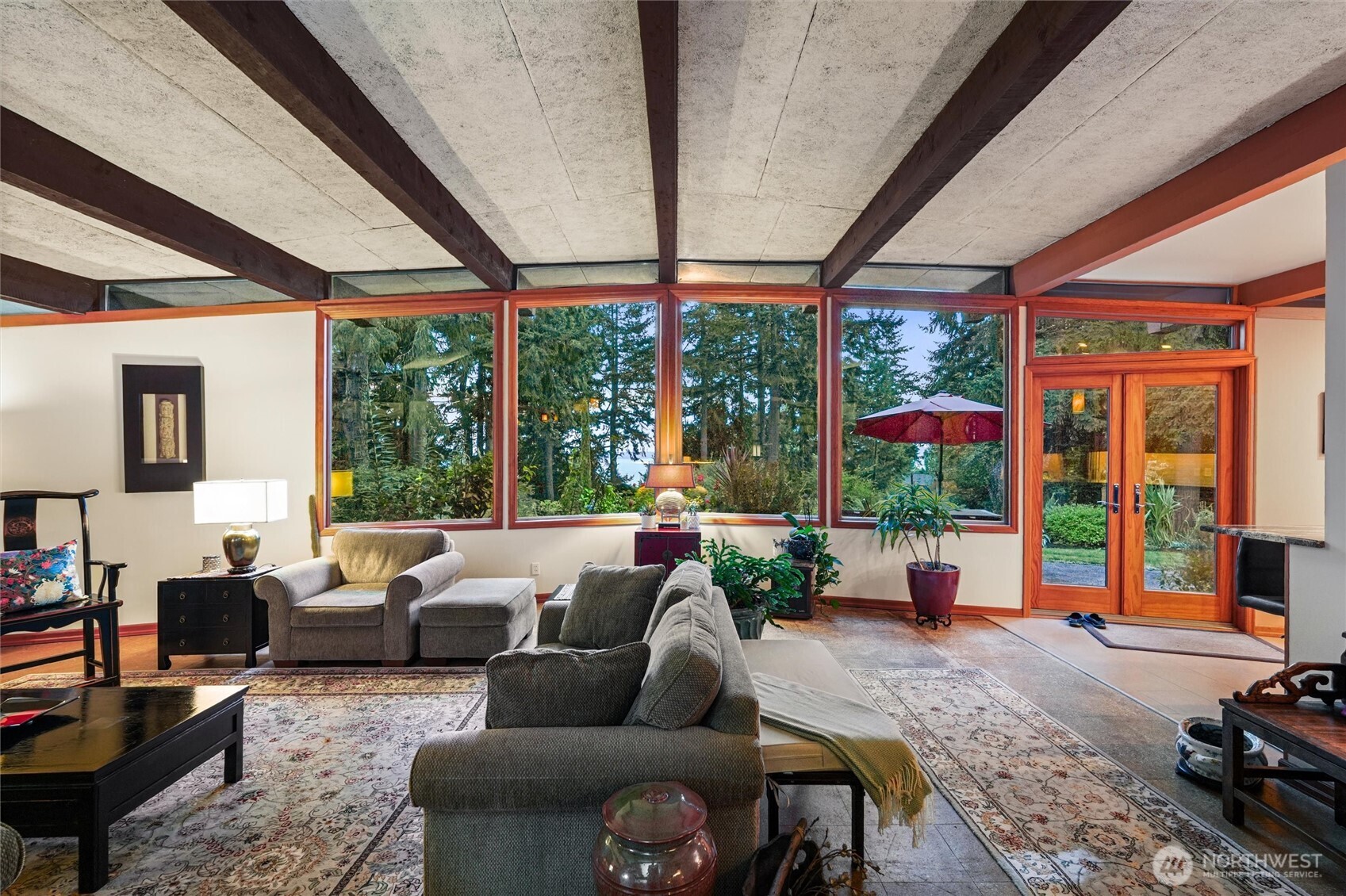 Furnished living room with exposed ceiling beams and large windows overlooking trees.