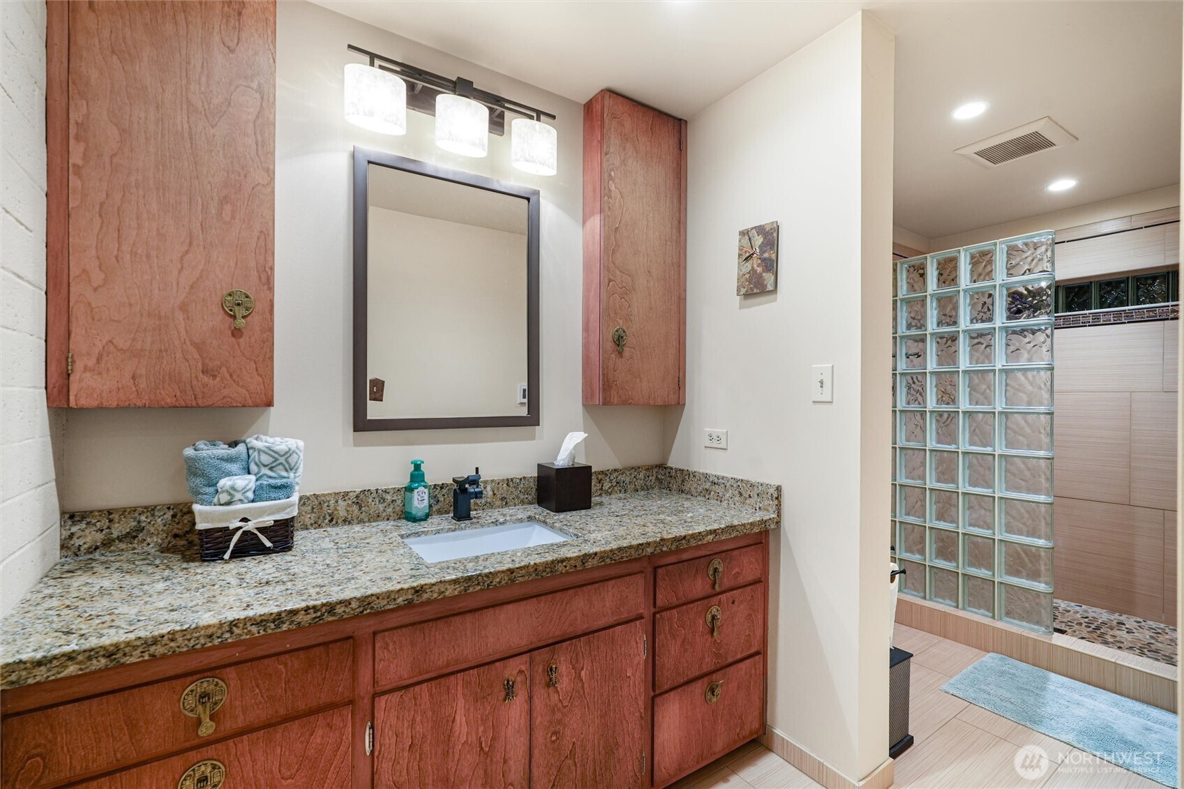 Bathroom with a granite vanity, reddish wood cabinetry, and a glass-block shower divider.