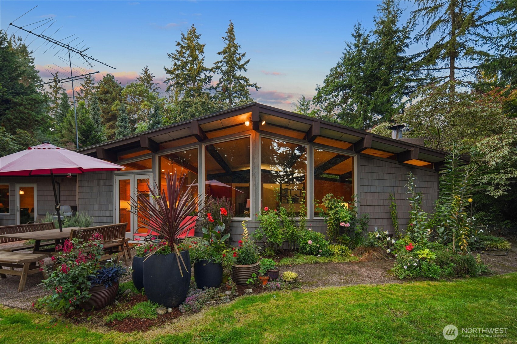 Midcentury-style home with a broad glass facade, landscaped garden, and outdoor dining area at dusk.