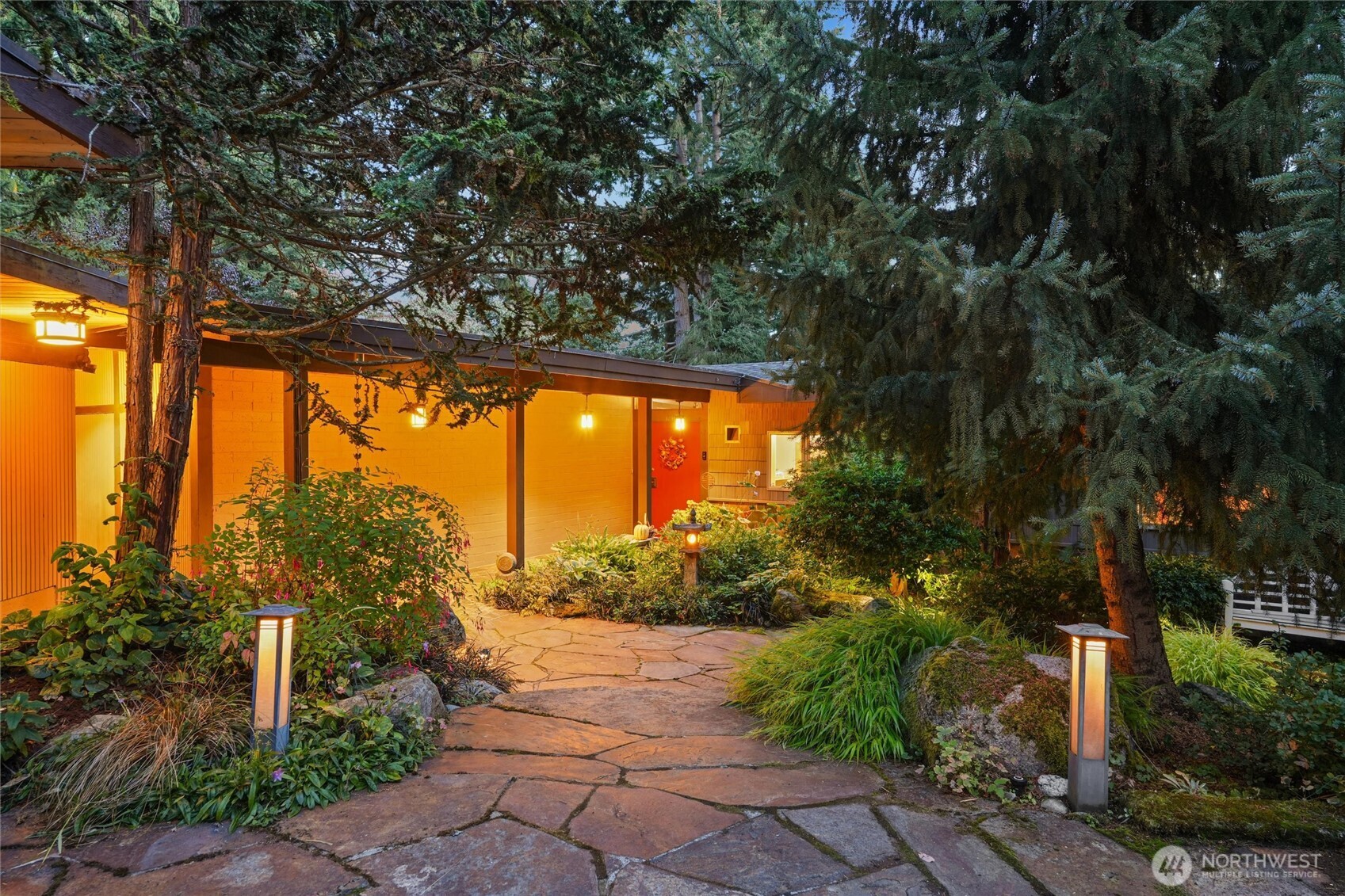 Single-story home's warmly lit entrance behind mature evergreens, with a flagstone walkway and garden lights.