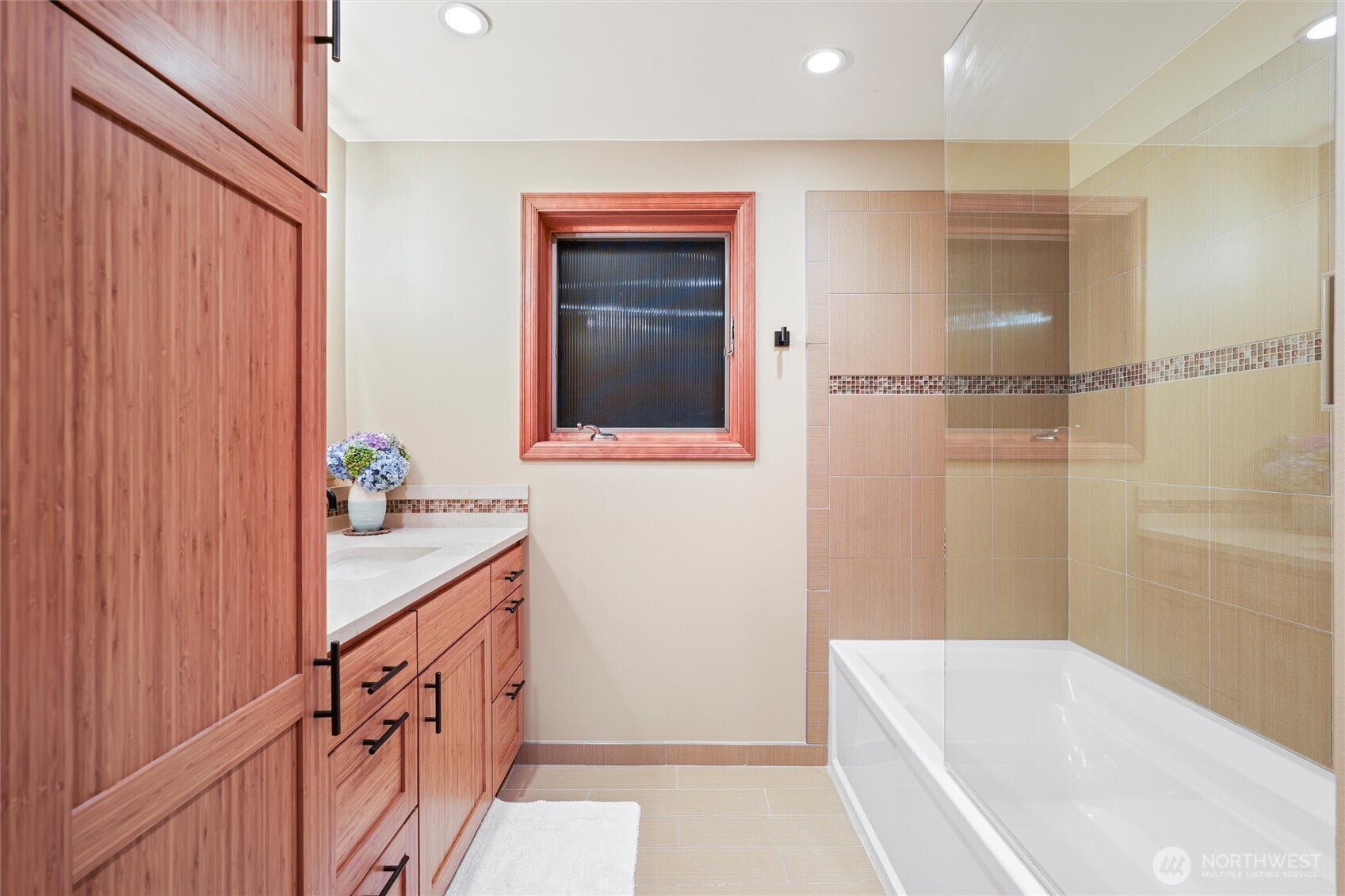 Bathroom with a glass-screened bathtub, beige tile, and reddish wood vanity.
