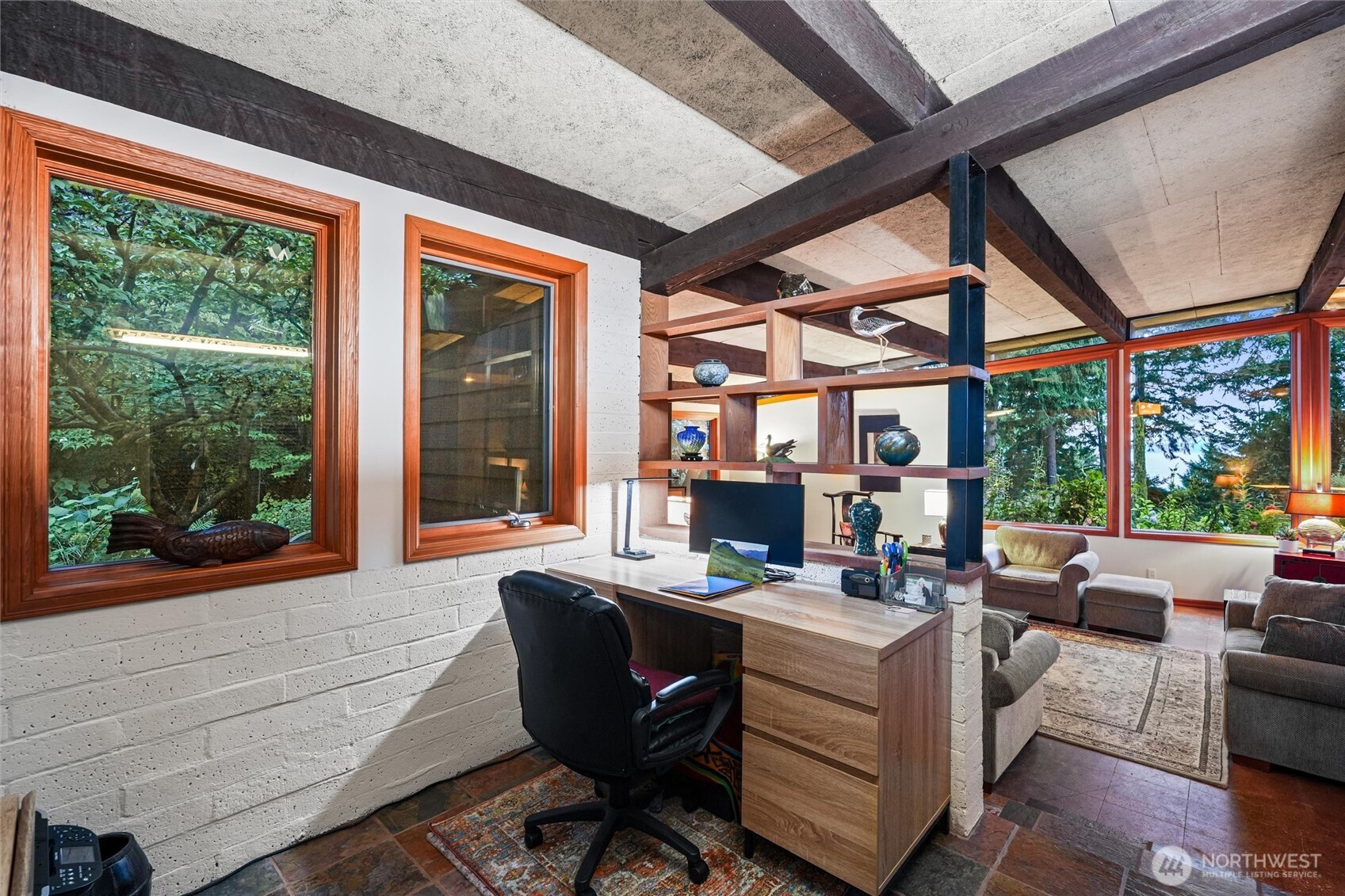 Home office with a desk and open shelving beside a living room with large windows overlooking trees.