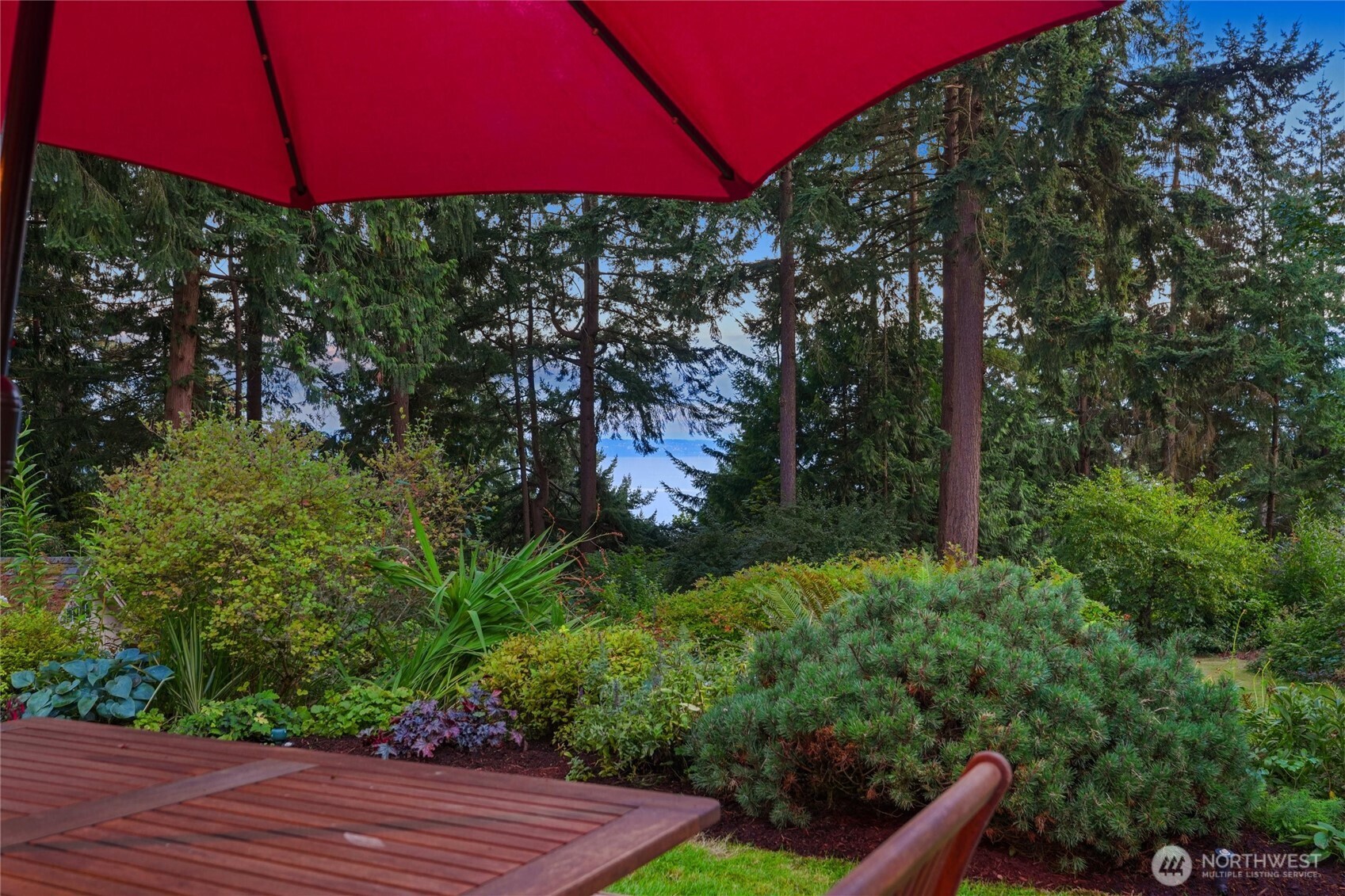 Wooden deck beneath a red umbrella overlooks a landscaped garden and water through evergreen trees.