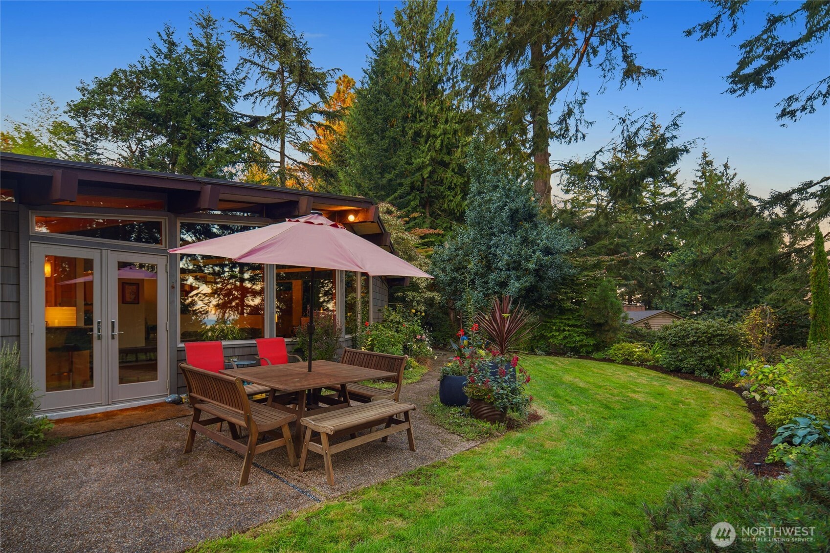 Backyard patio with a wooden dining set and pink umbrella beside large glass doors and a landscaped lawn.