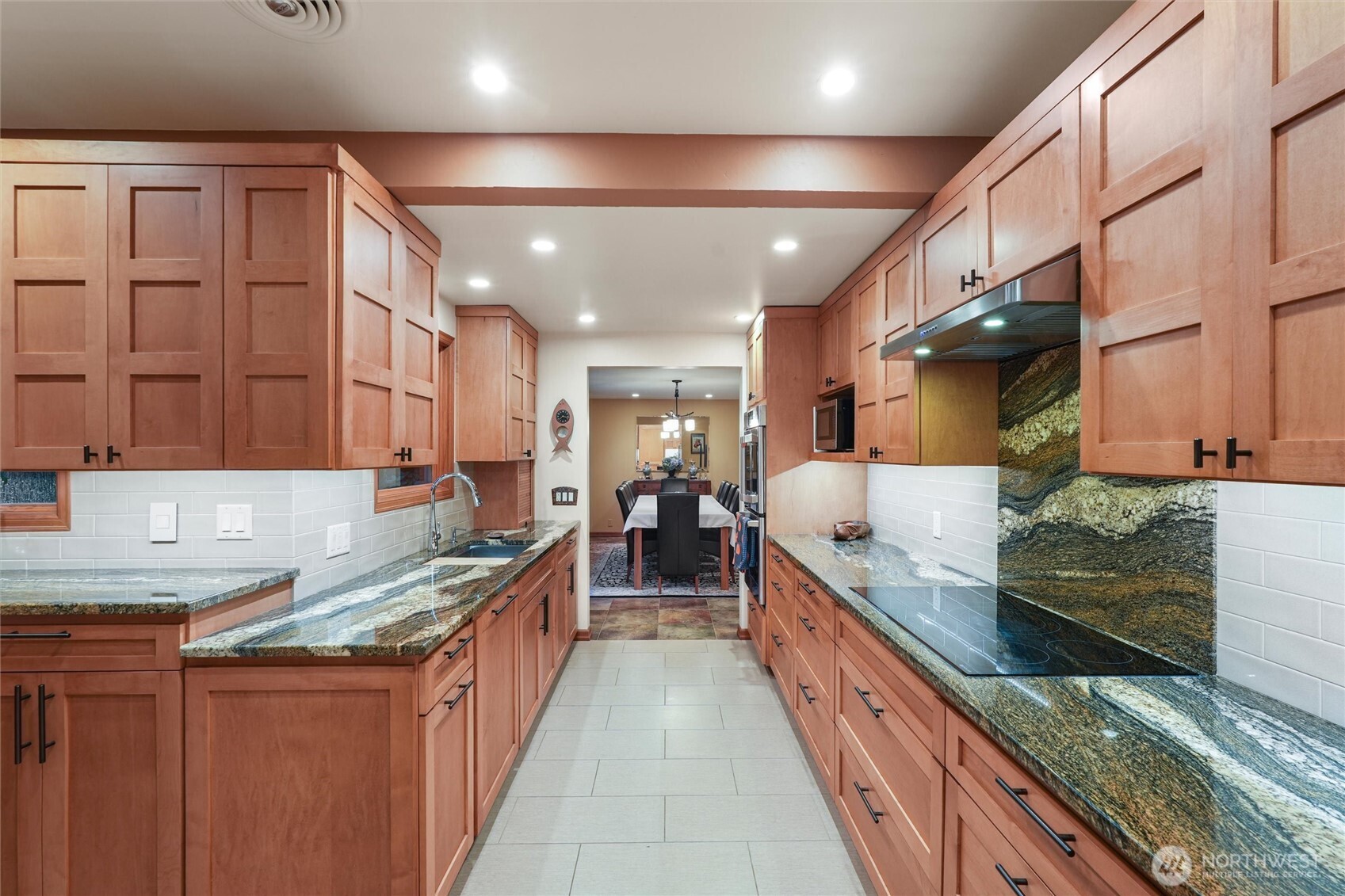 Galley kitchen with reddish wood cabinets, granite counters, and a cooktop beneath a stainless range hood.