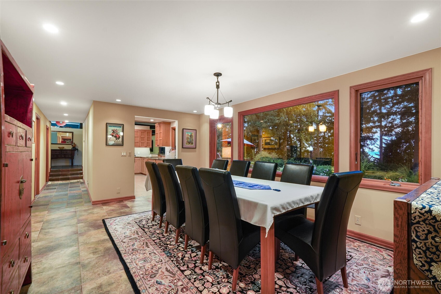 Dining room with a long table, black chairs, patterned rug, and large windows overlooking trees.