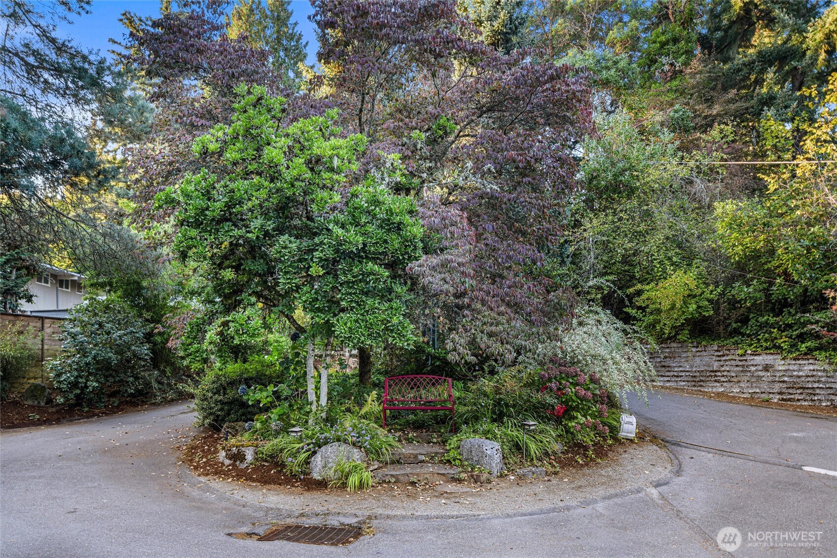 Landscaped garden with a red bench, mature trees, and a paved road curving around it.