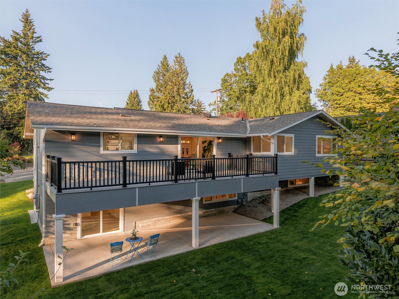Gray home with a raised black-railed deck, covered patio, and mature trees in warm evening light.
