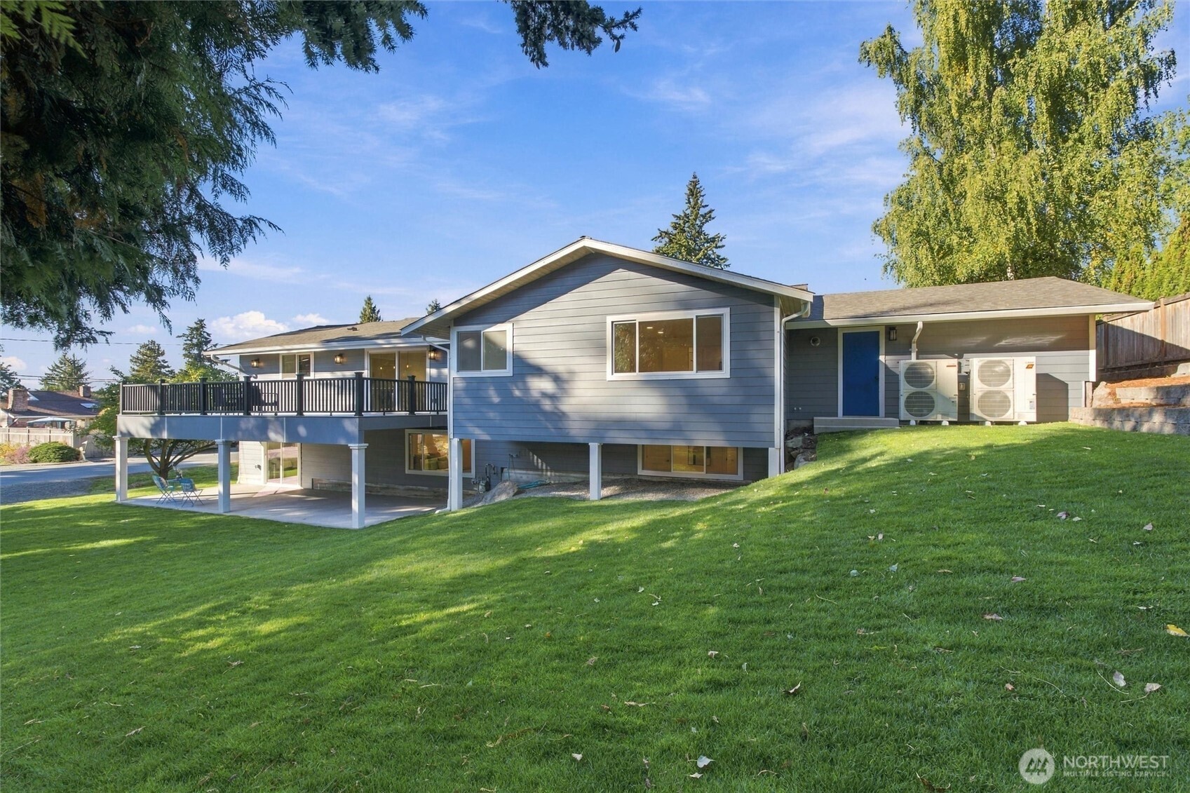 Rear exterior of a blue-gray house with a raised deck, covered patio, and sloping lawn.