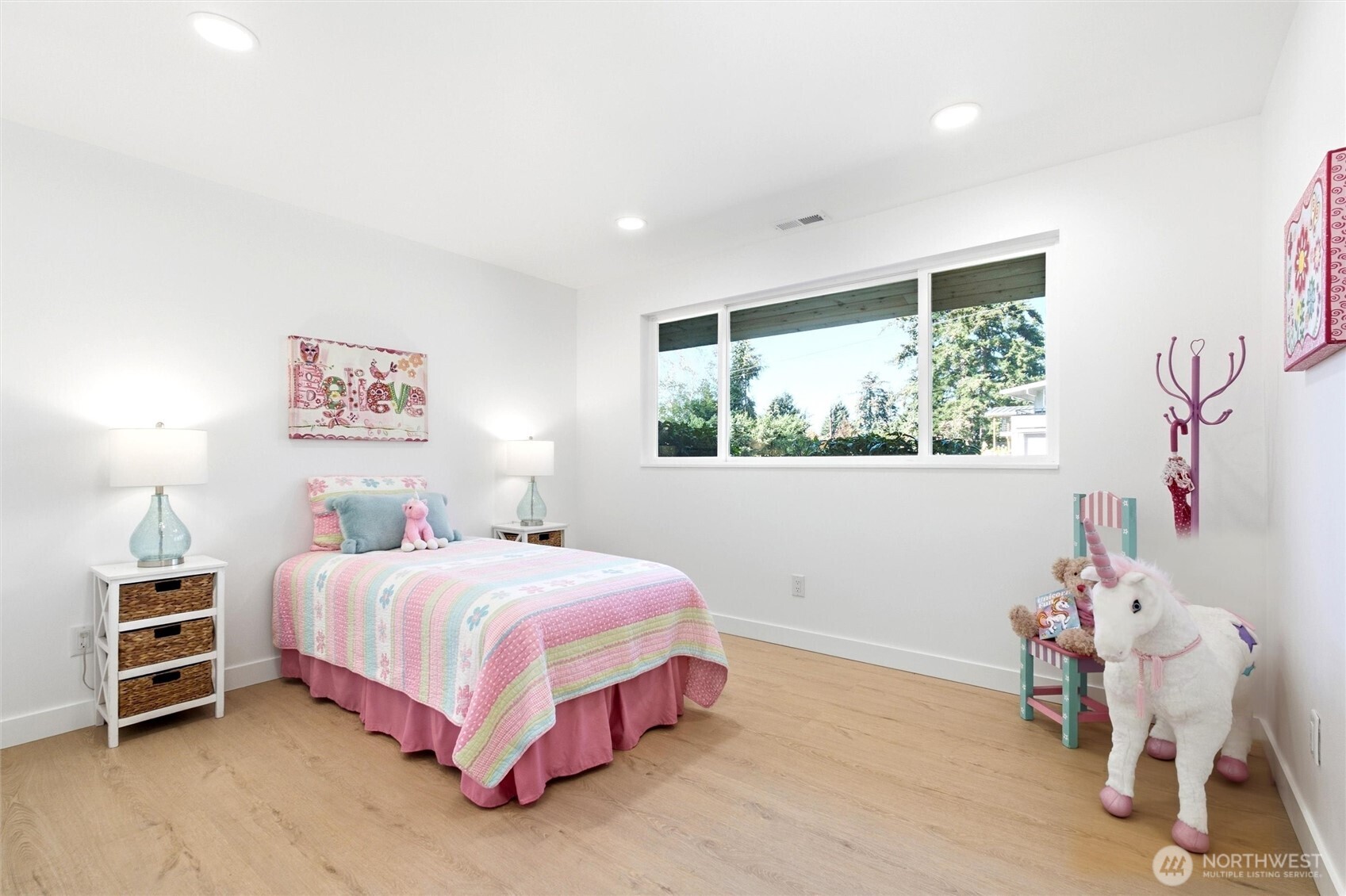 Bedroom with pastel quilted bed, large window overlooking greenery, and a plush unicorn.