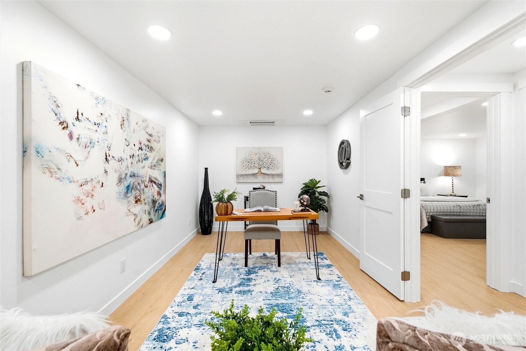 Home office with a wood desk, blue-and-white rug, and large abstract artwork; bedroom visible through the open doorway.