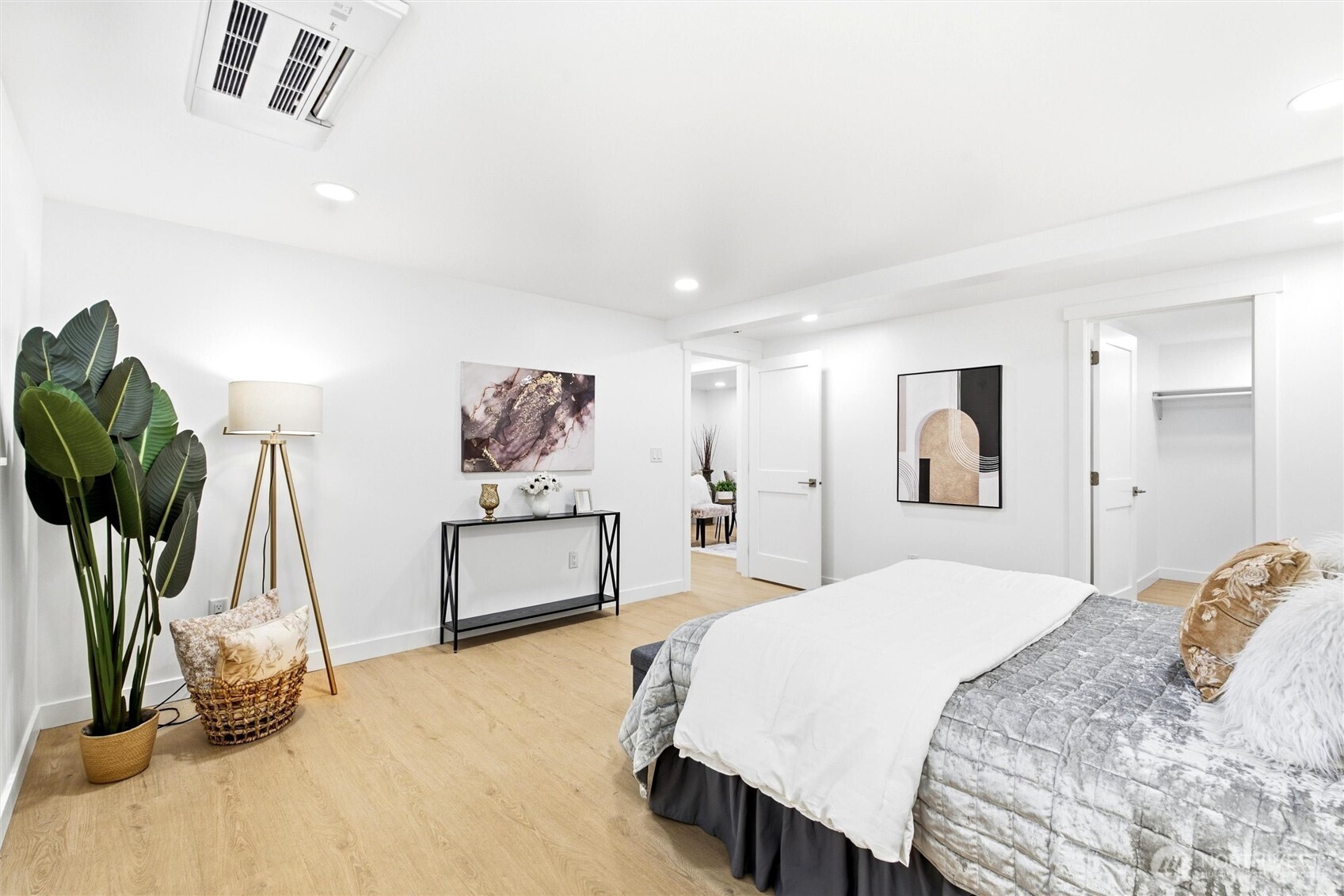 Staged bedroom with white and gray bedding, light wood floors, and a large potted plant.