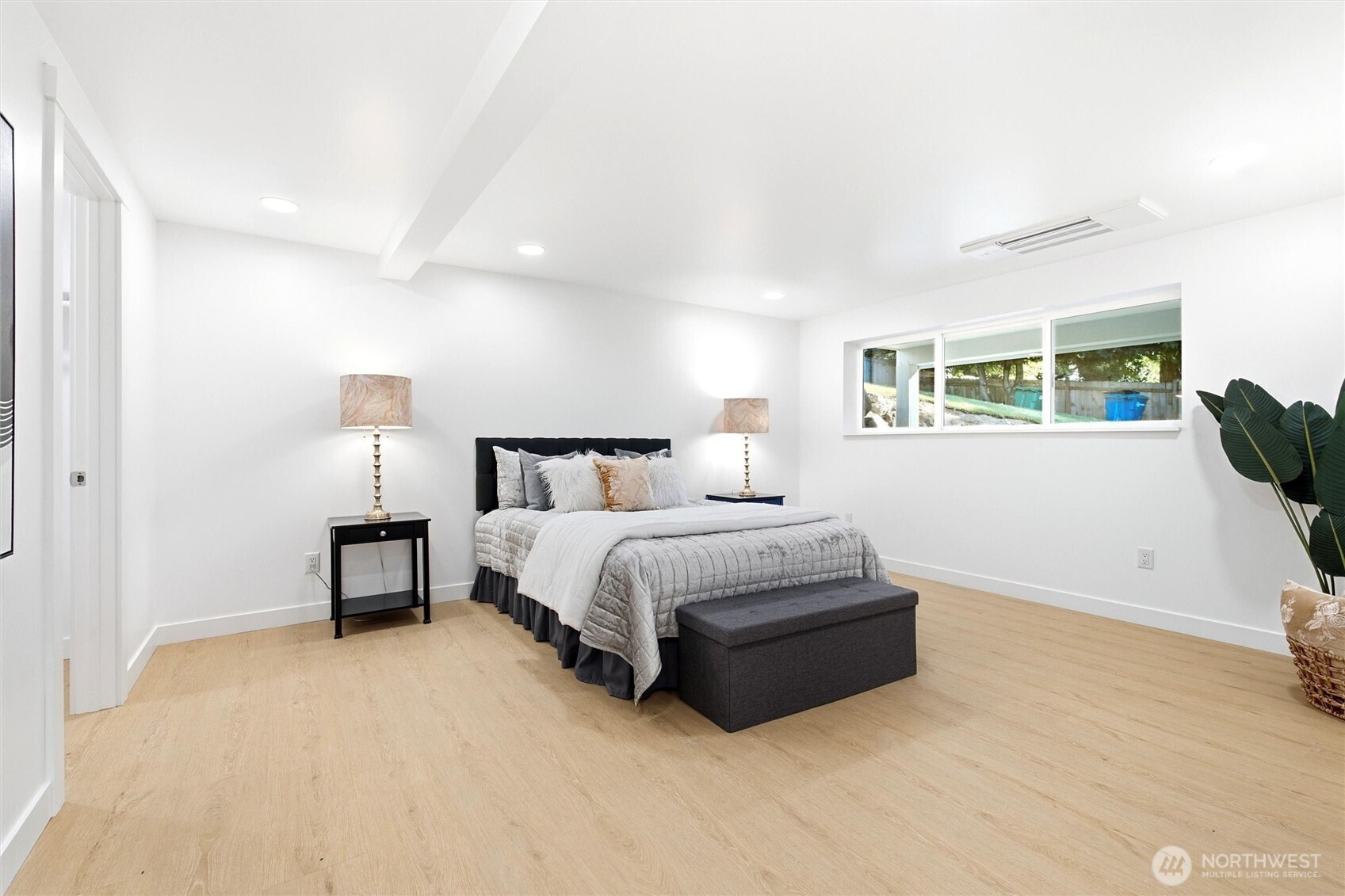 Staged bedroom with a gray bed, dark bench, light wood flooring, and a window overlooking the yard.