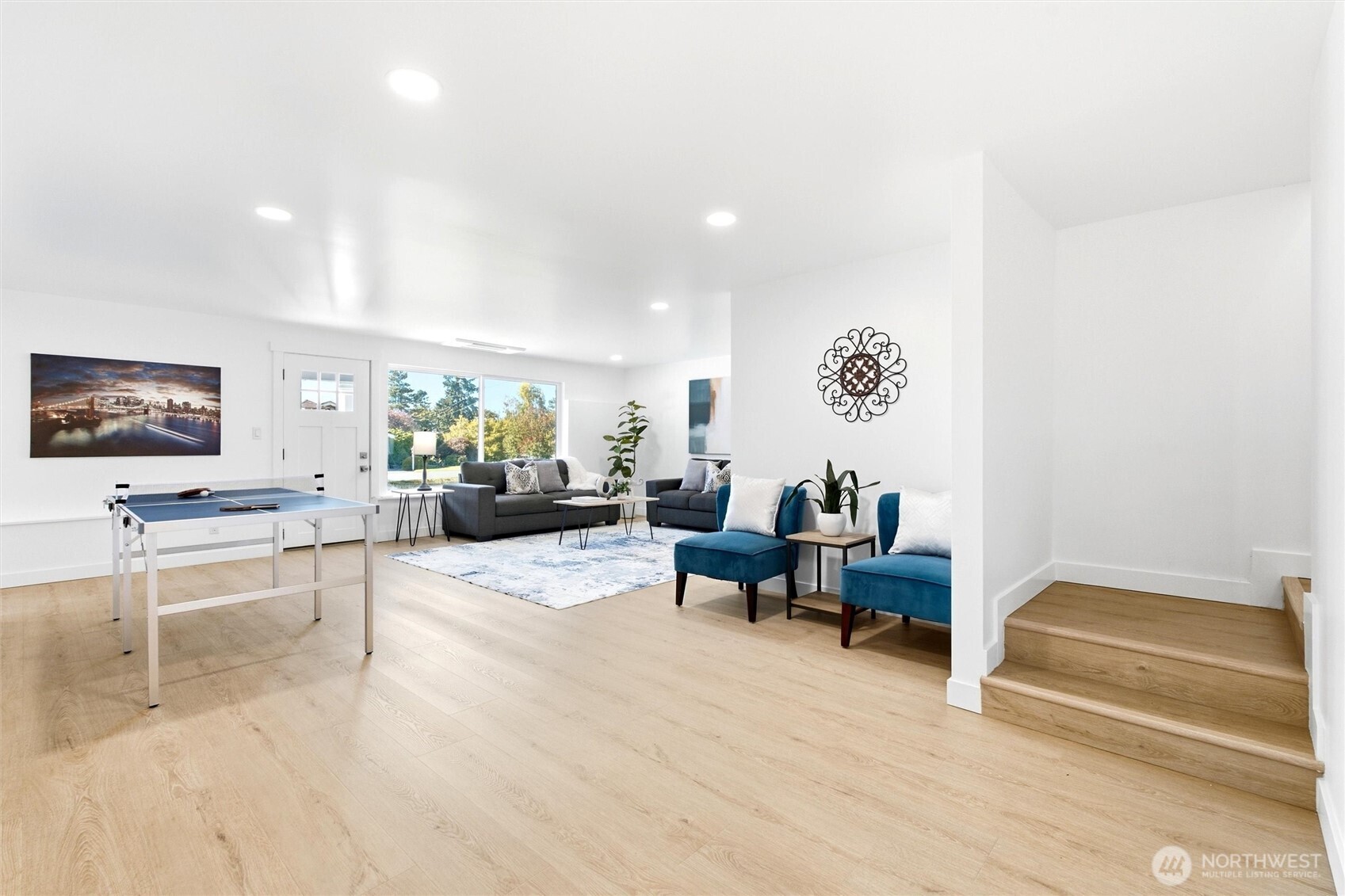Furnished recreation room with a table tennis table, gray sofas, teal chairs, and wood stairs.