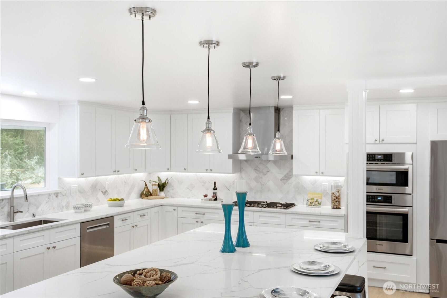 Bright white kitchen with a large veined-stone island, glass pendants, and stainless steel appliances.