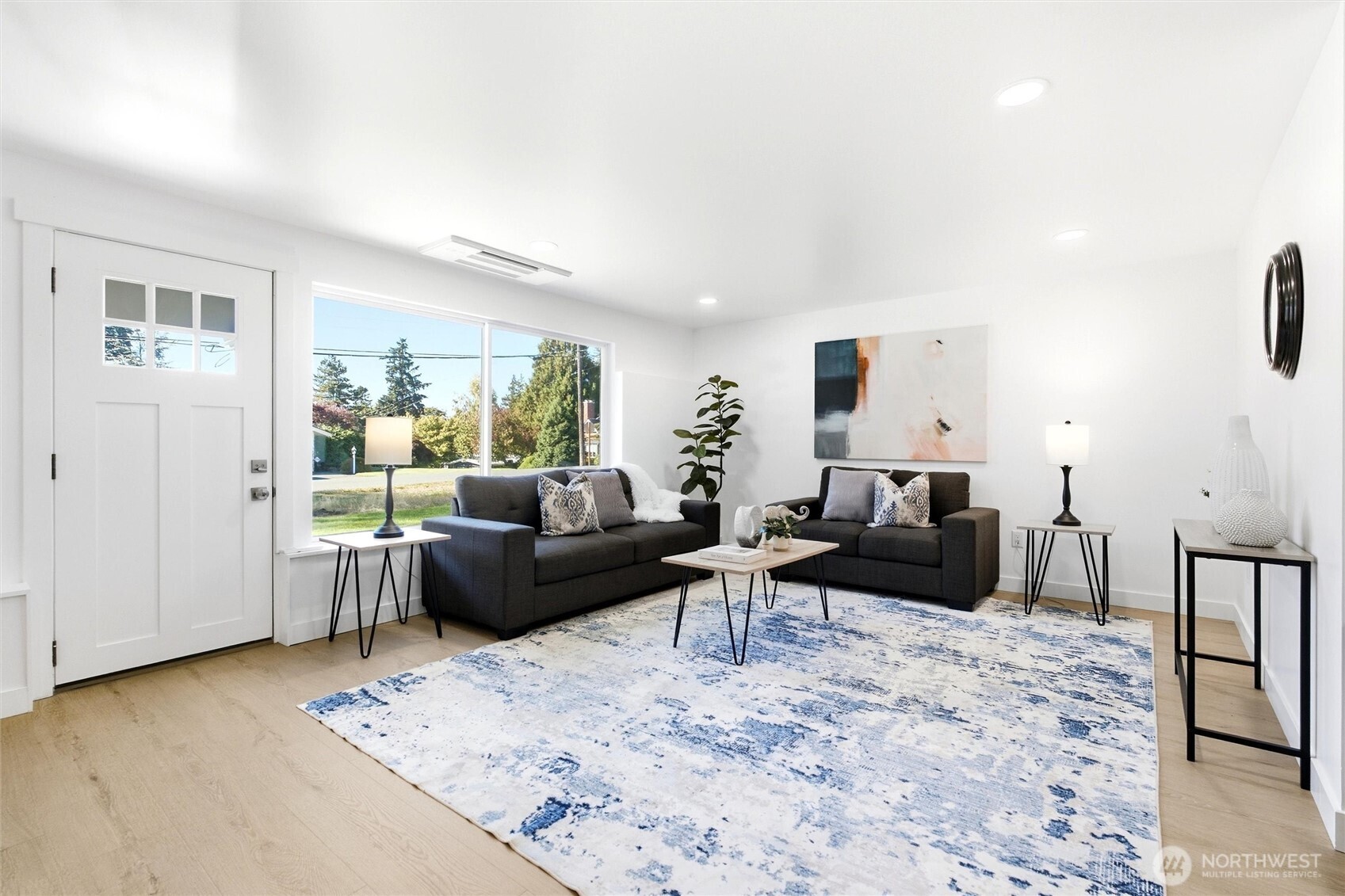 Staged living room with charcoal sofas, a blue-and-white rug, and large windows beside the front door.