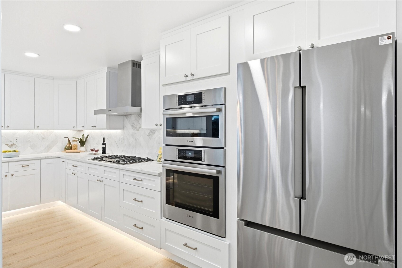 White kitchen with marble backsplash, gas cooktop, stacked ovens, and stainless-steel refrigerator.