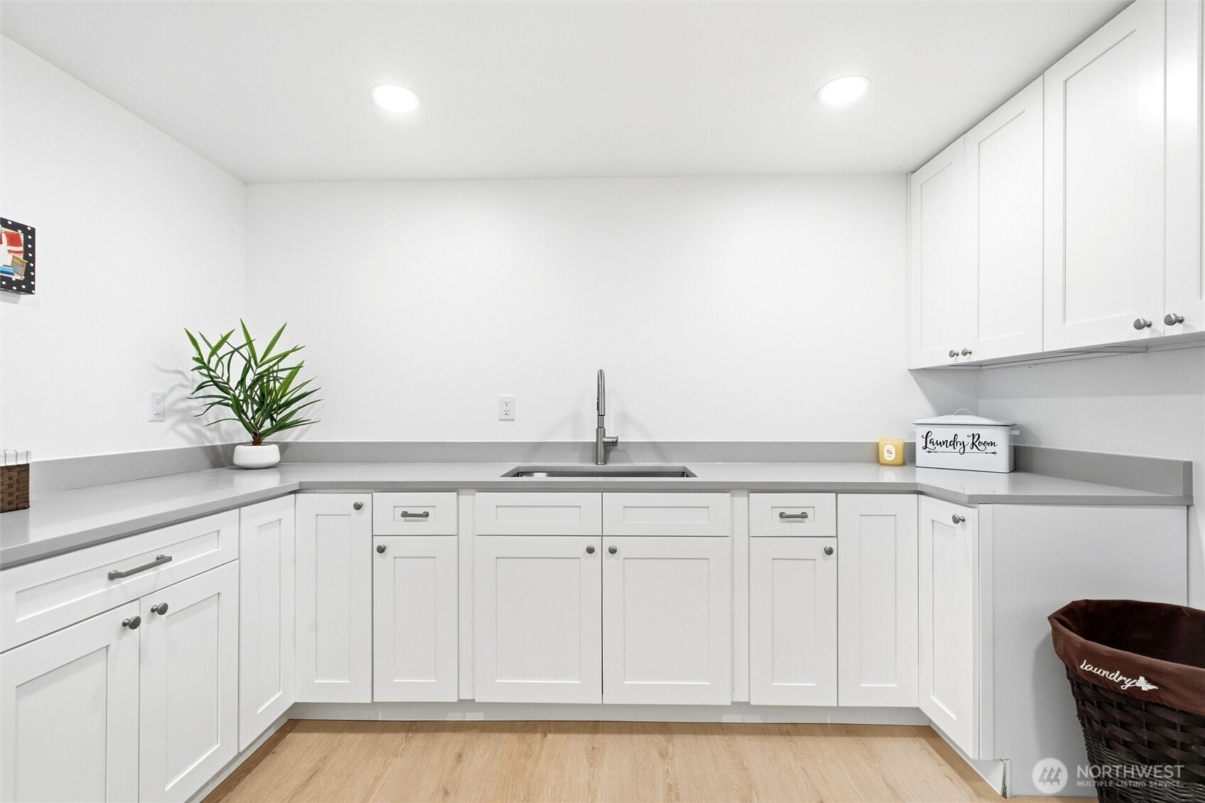 Laundry room with white cabinets, gray counters, a central sink, and light wood flooring.