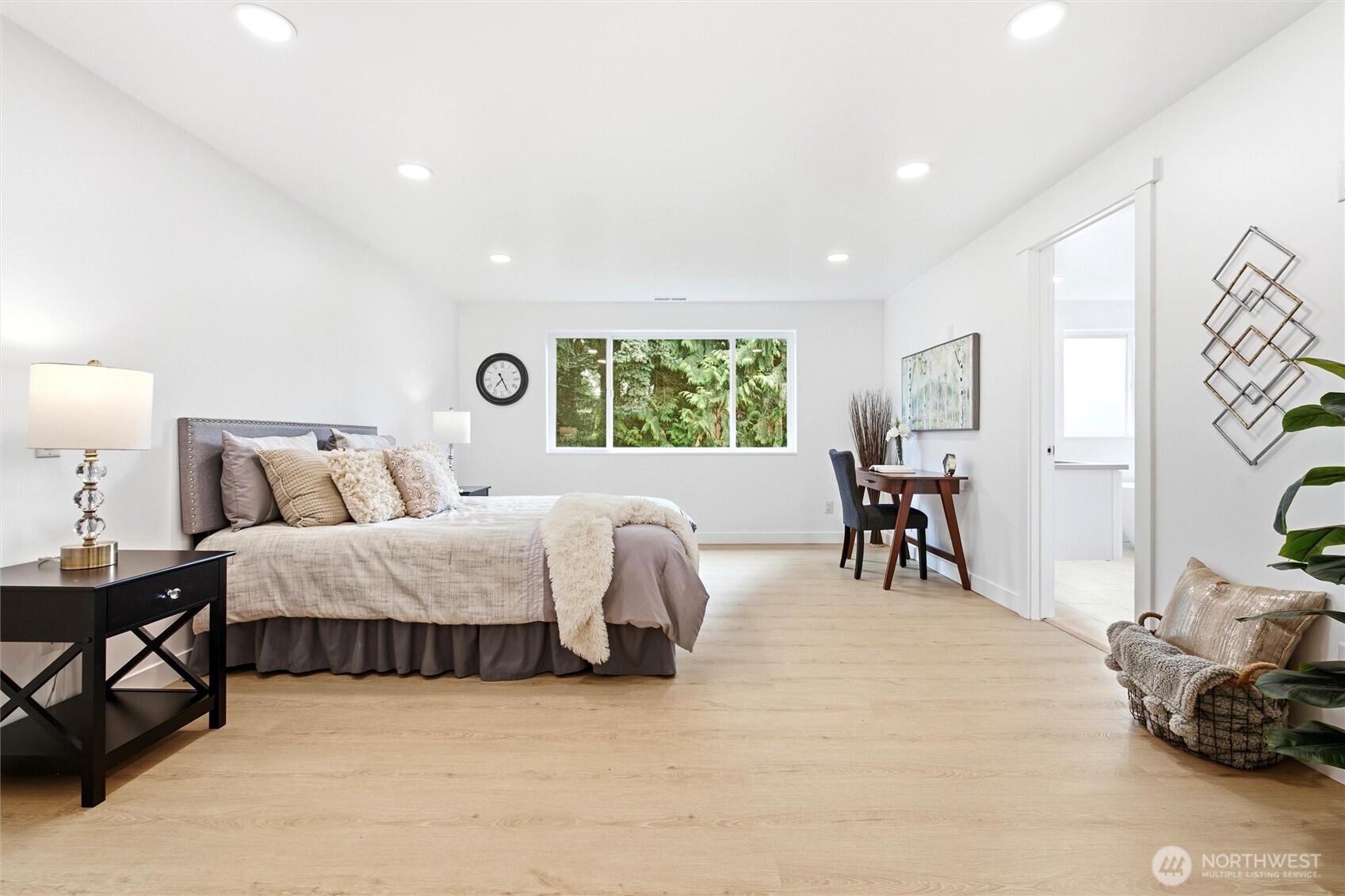 Staged bedroom with a neutral bed, light wood floors, and a window overlooking greenery.