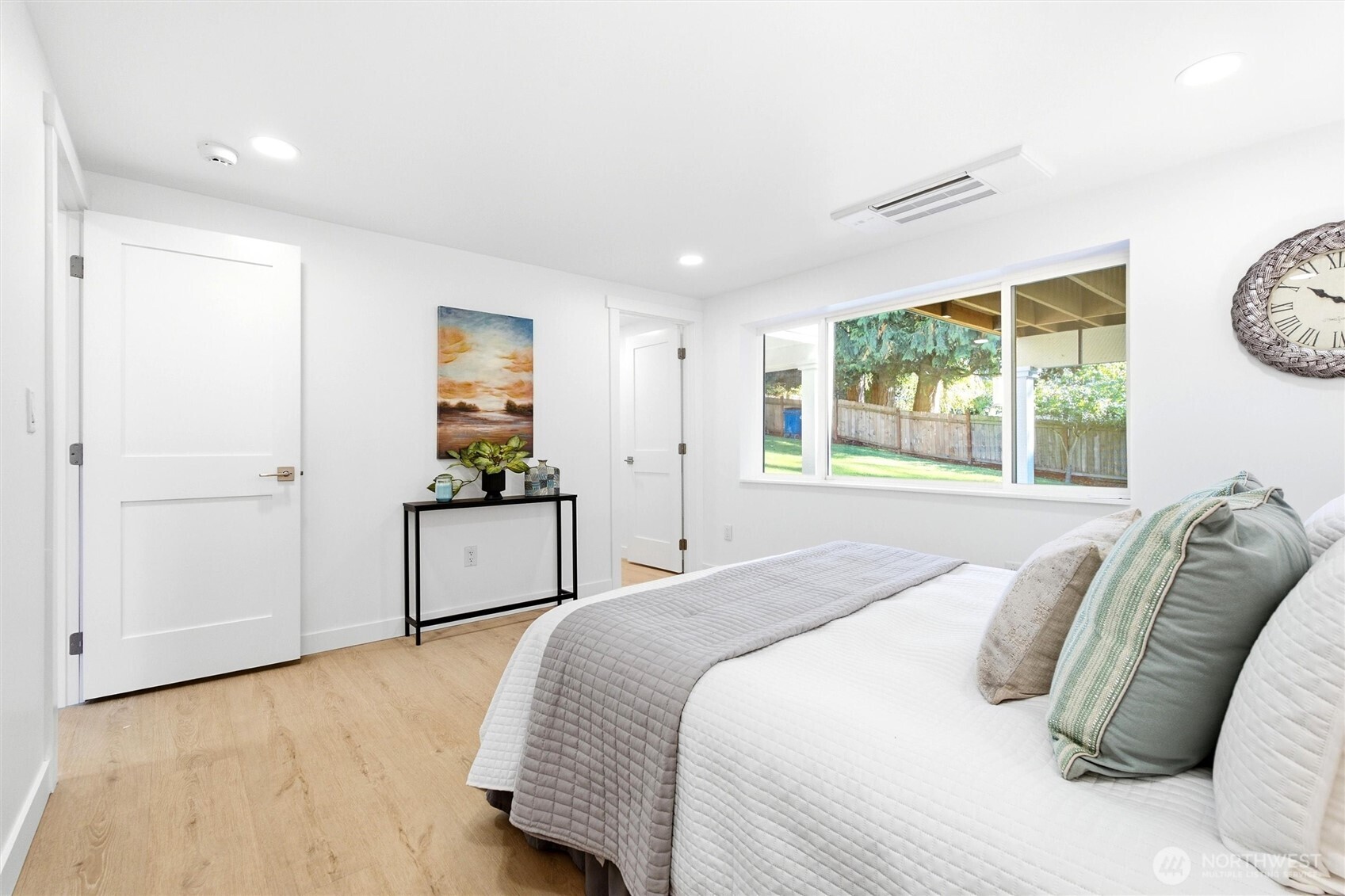 Furnished bedroom with a white bed, gray quilt, and wide window overlooking a fenced backyard.