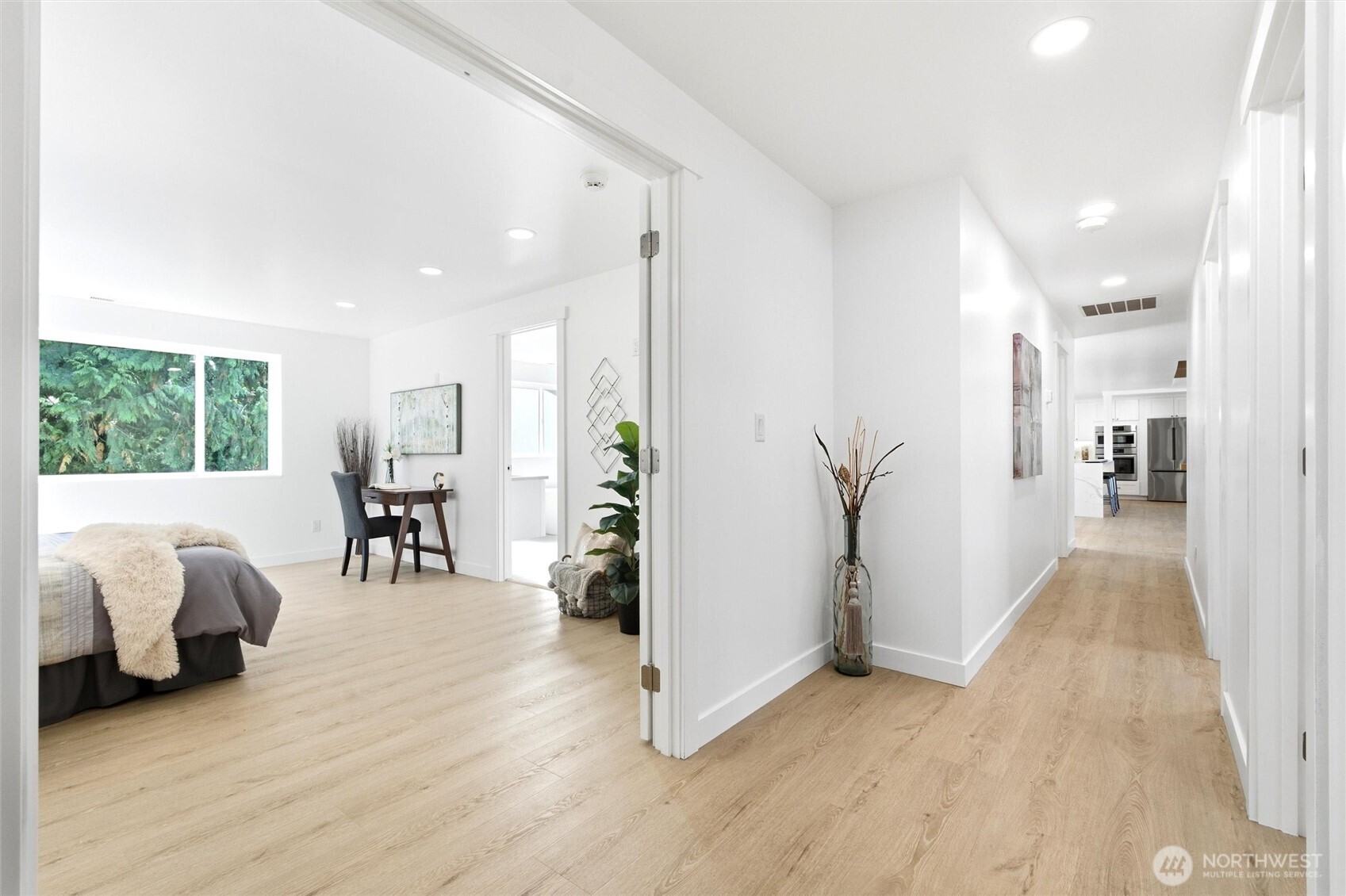 Bright hallway with light wood floors, opening onto a furnished bedroom and a distant kitchen.
