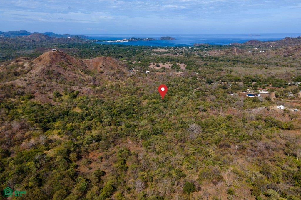 Aerial view of wooded hills marked by a red pin, with the ocean visible in the distance.