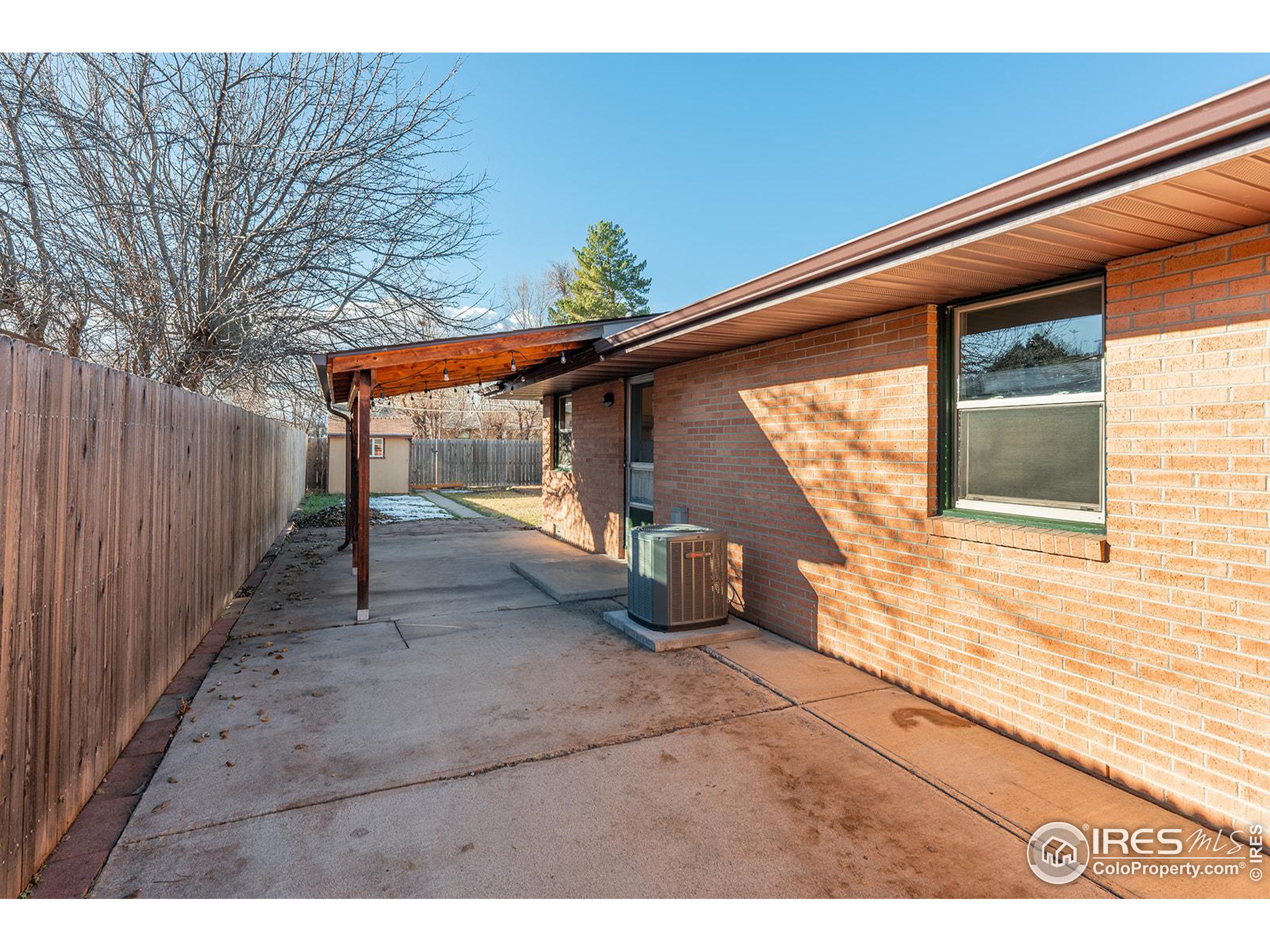 Covered concrete side patio beside a brick house, with a wooden privacy fence and air-conditioning unit.