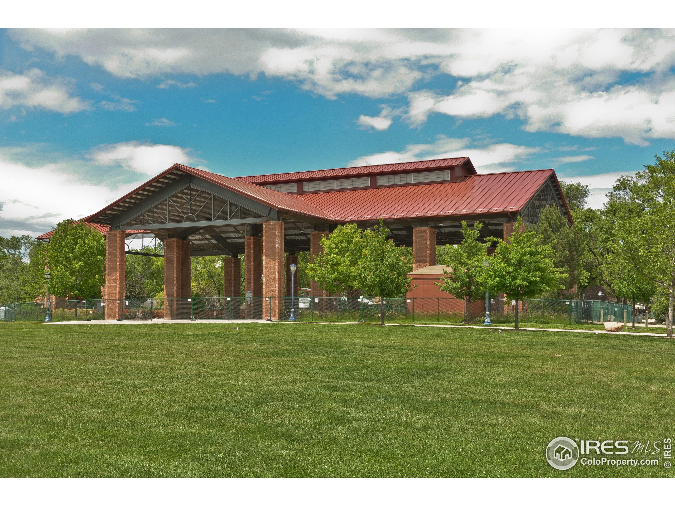Open-sided pavilion with a red metal roof and brick columns beside a green lawn.