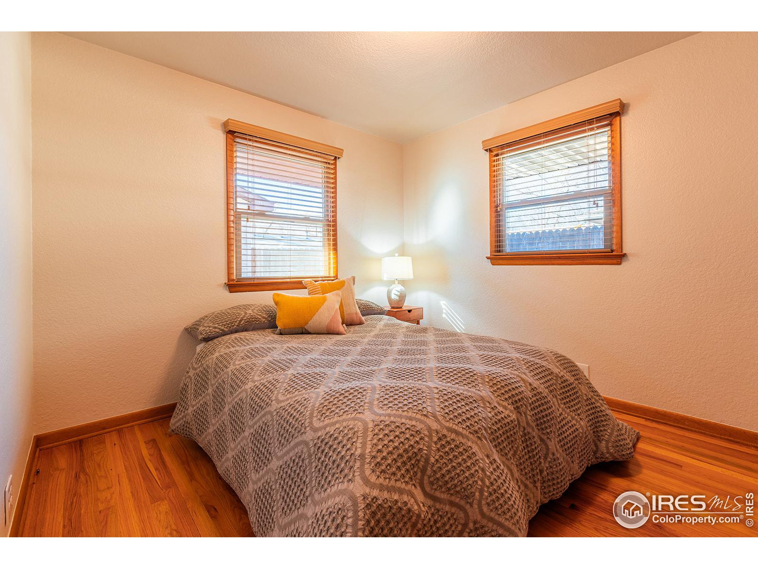Furnished bedroom with a patterned bedspread, two wood-trimmed windows, and hardwood floors.