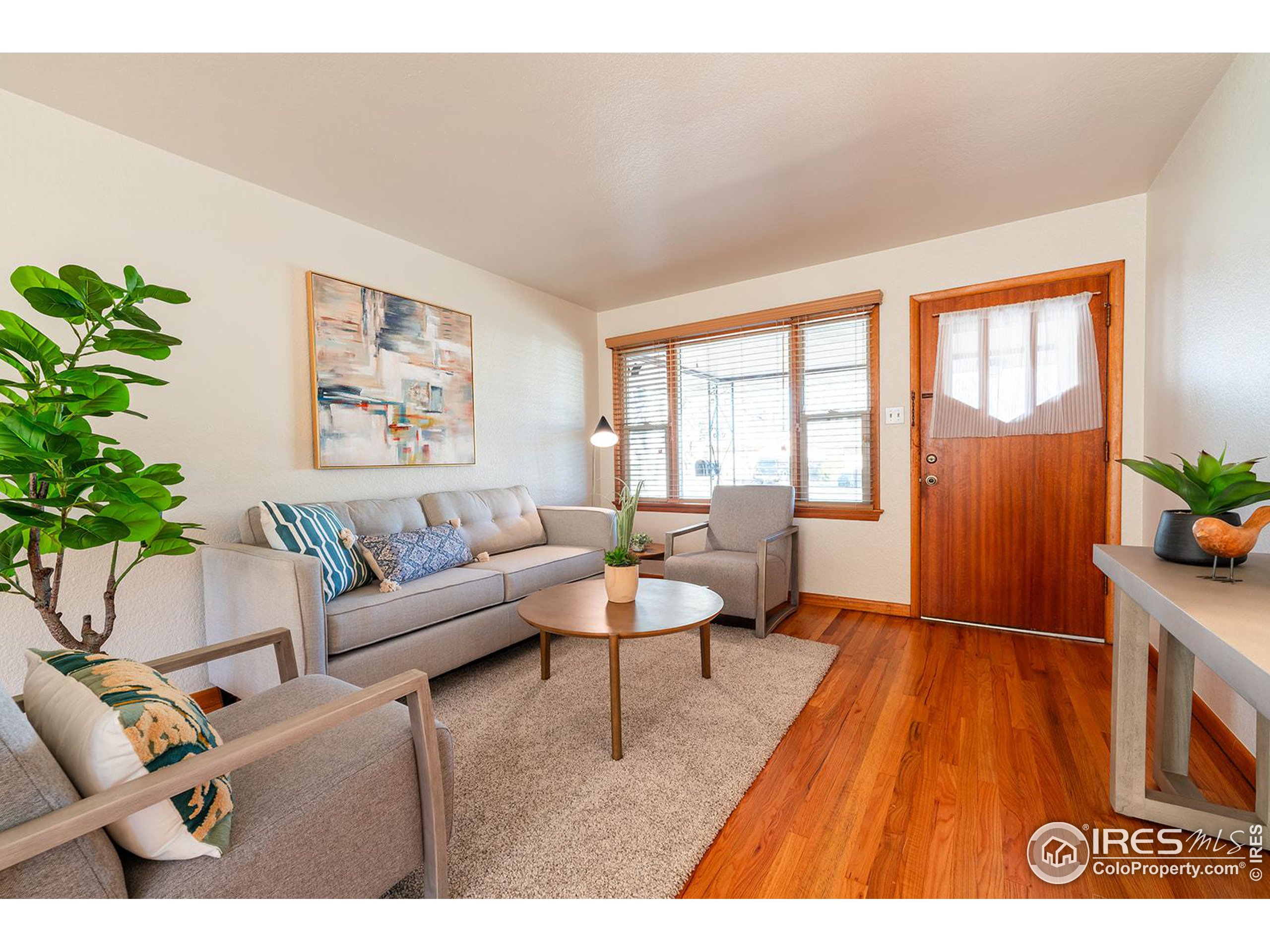 Staged living room with gray seating, hardwood floors, and a wood-framed entry door.