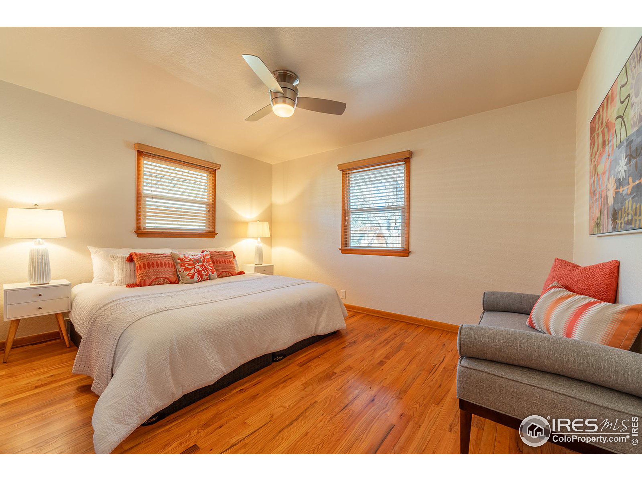 Bedroom with a white-covered bed, coral pillows, hardwood floors, and a gray chair.