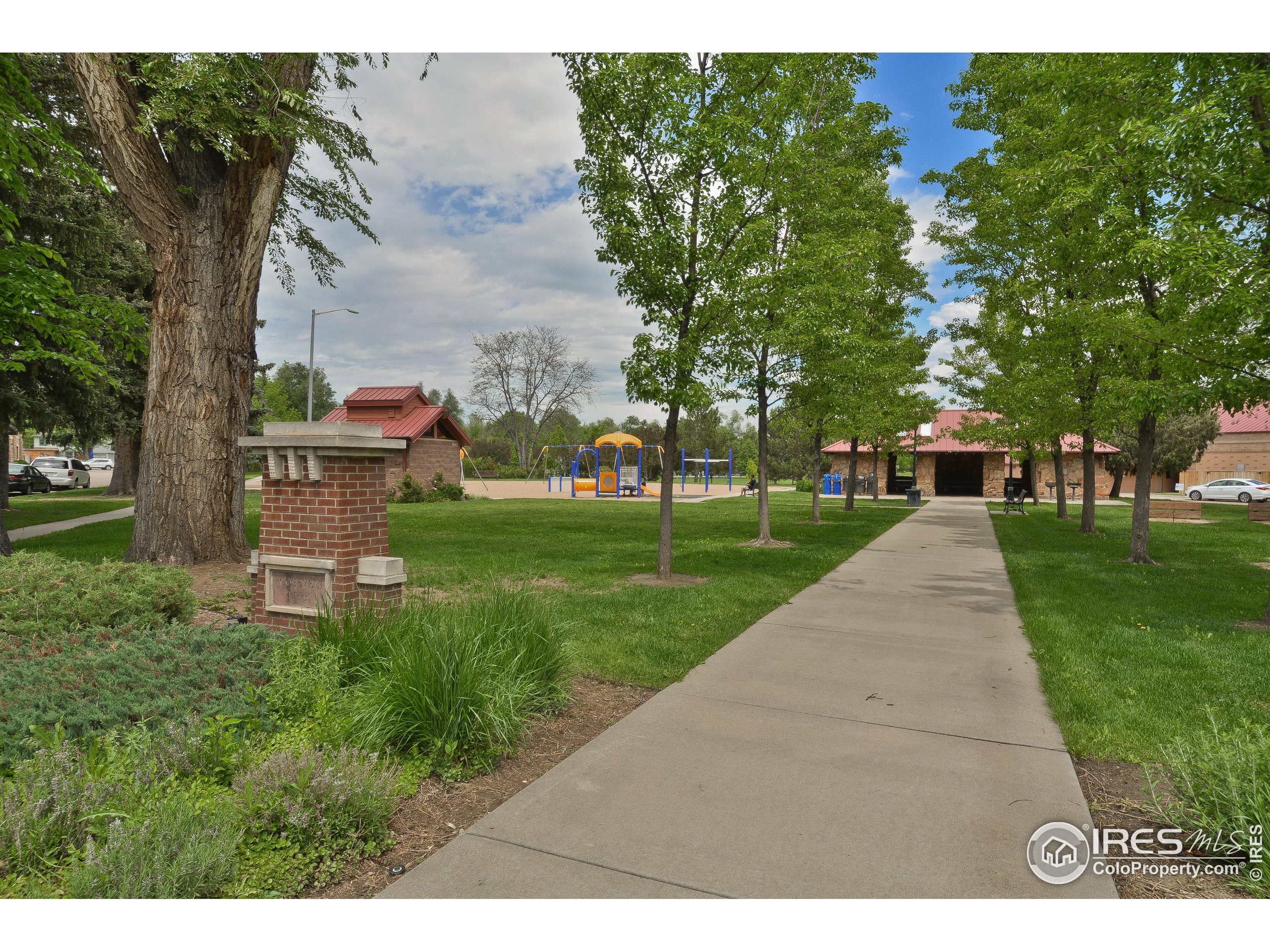 Neighborhood park with a playground, red-roofed pavilion, and paved path beneath mature trees.
