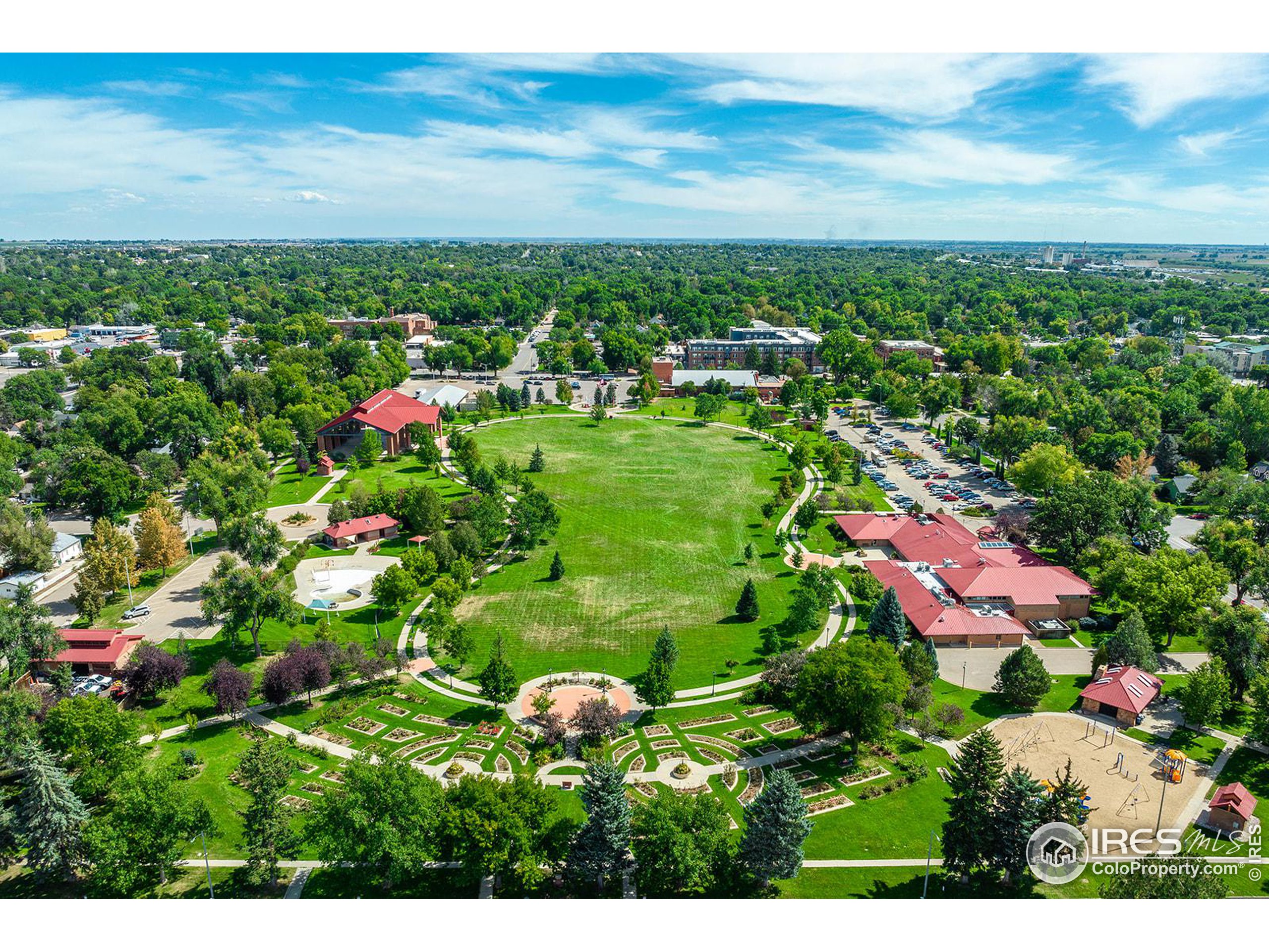 Aerial view of a leafy park with a broad lawn, winding paths, formal gardens, and a playground.