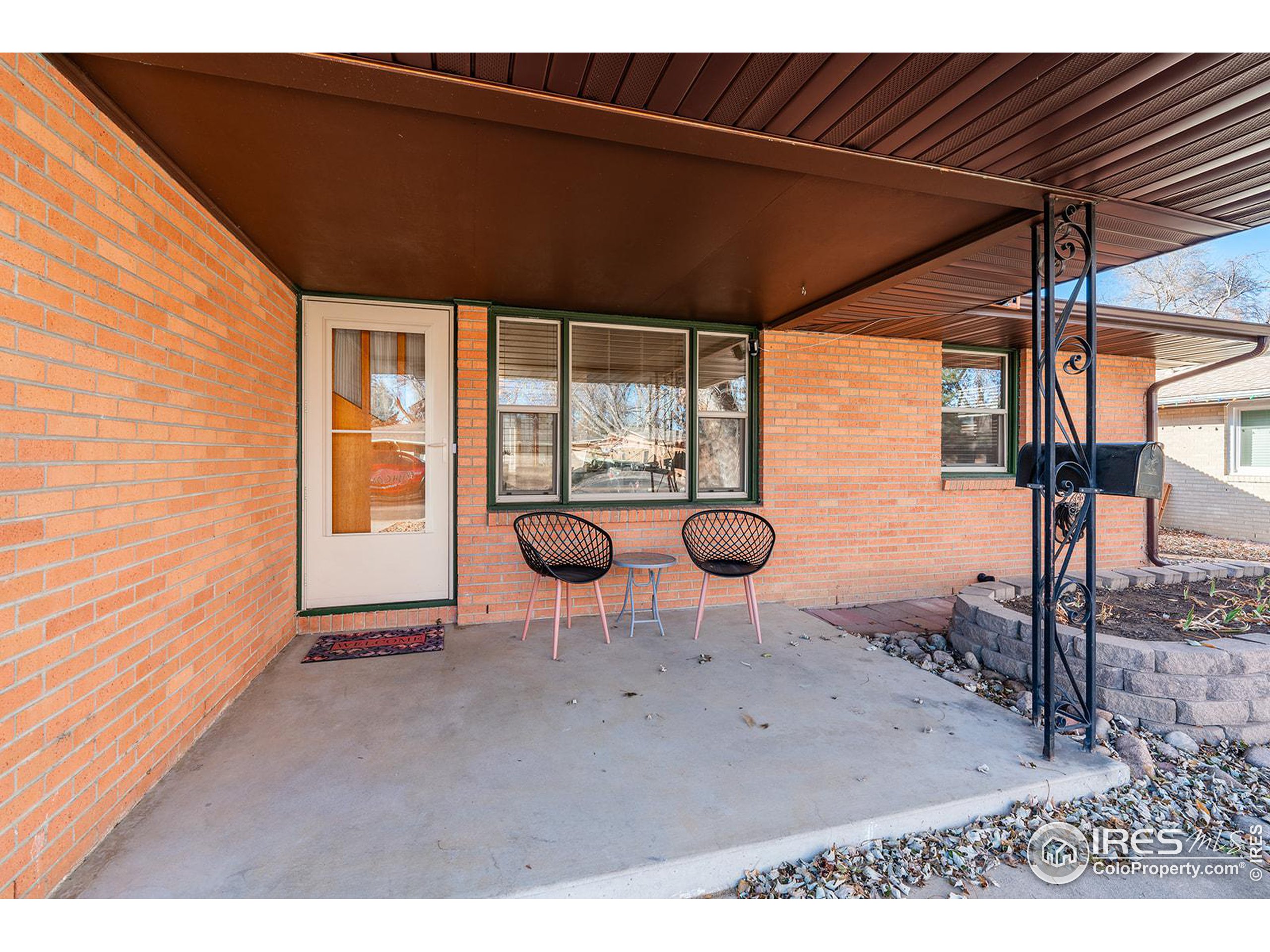 Brick home’s covered front porch with two black chairs, a small table, and decorative metal supports.
