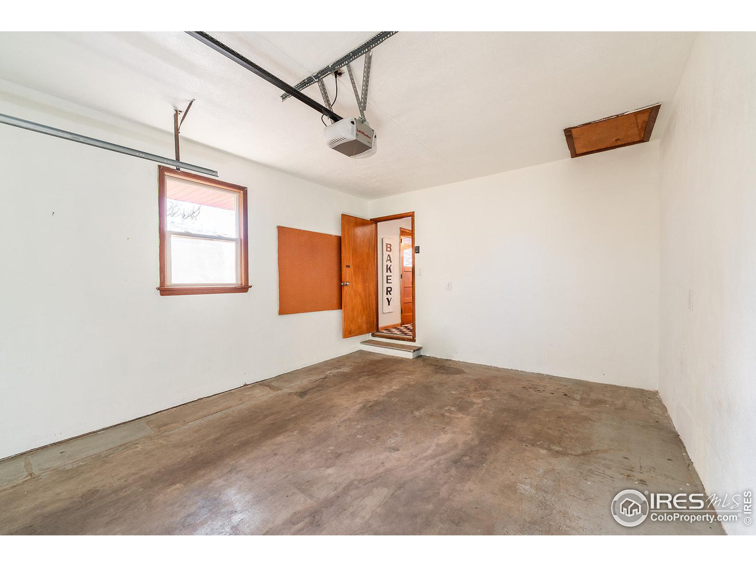 Empty garage with worn concrete floor, window, overhead door tracks, and doorway marked “BAKERY.”