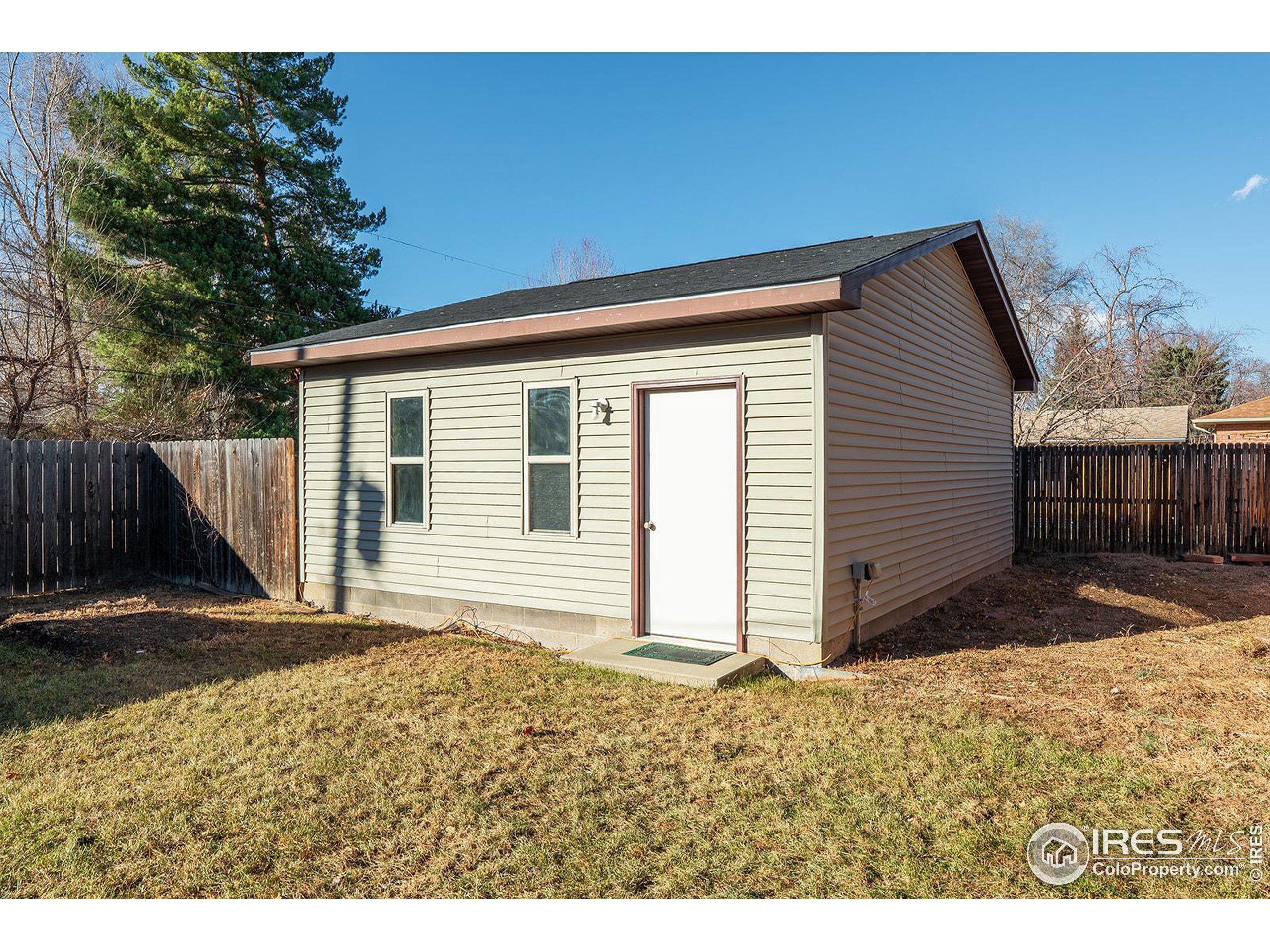 Detached backyard outbuilding with two narrow windows and a white door, enclosed by a wooden fence.