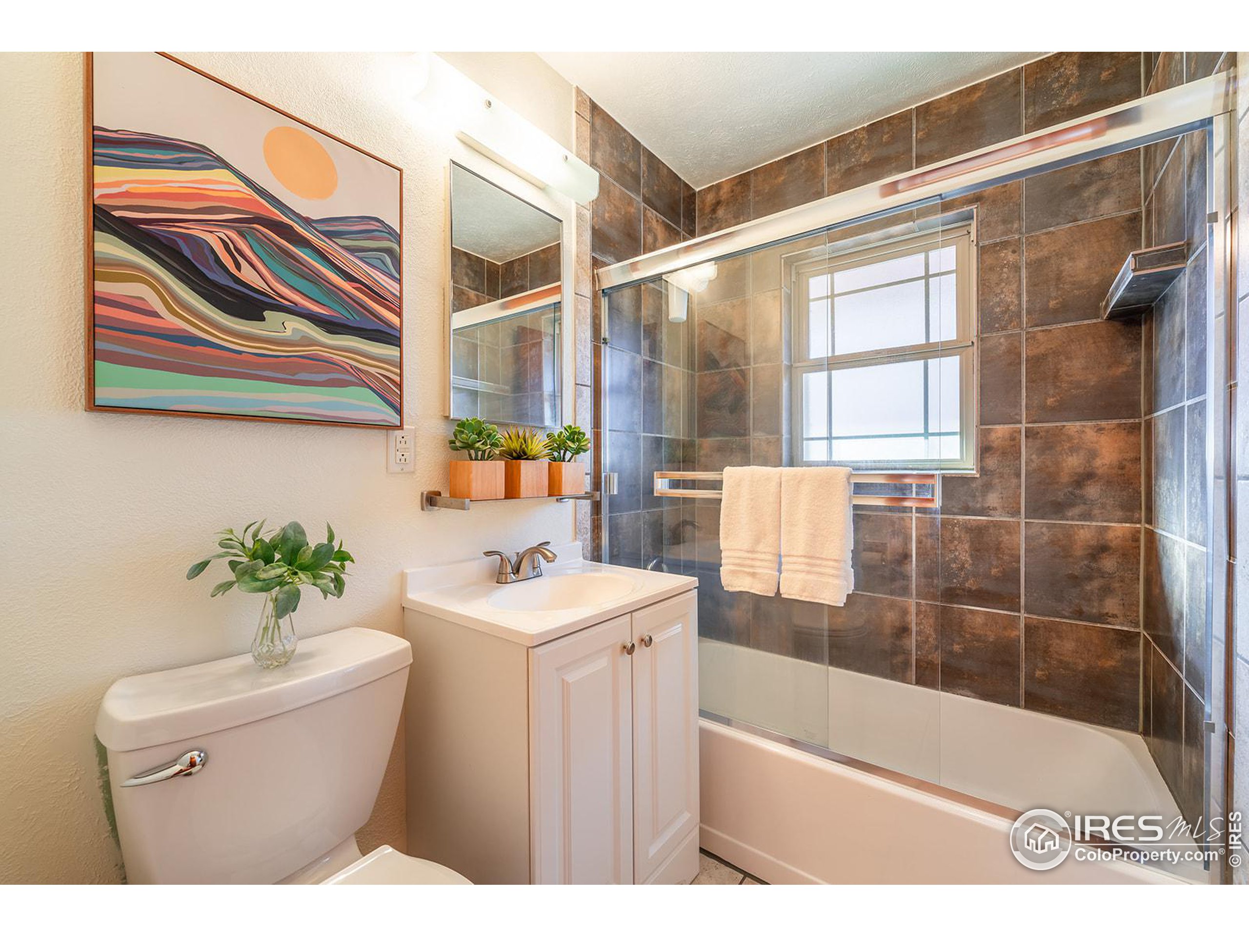 Bathroom with a glass-enclosed tub, dark tile, white vanity, and colorful framed artwork.
