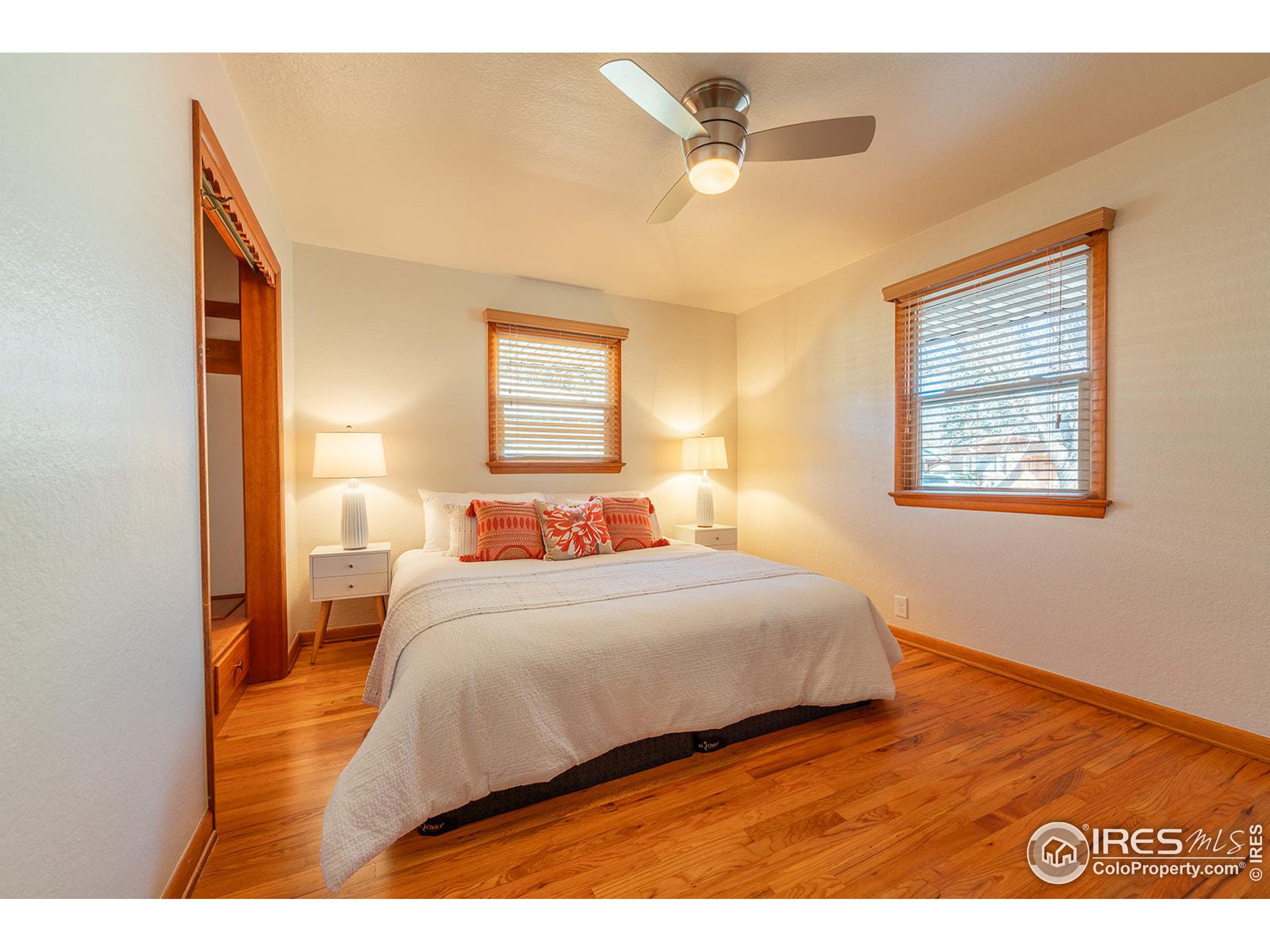 Furnished bedroom with a white bed, coral pillows, hardwood floors, and wood-trimmed windows.
