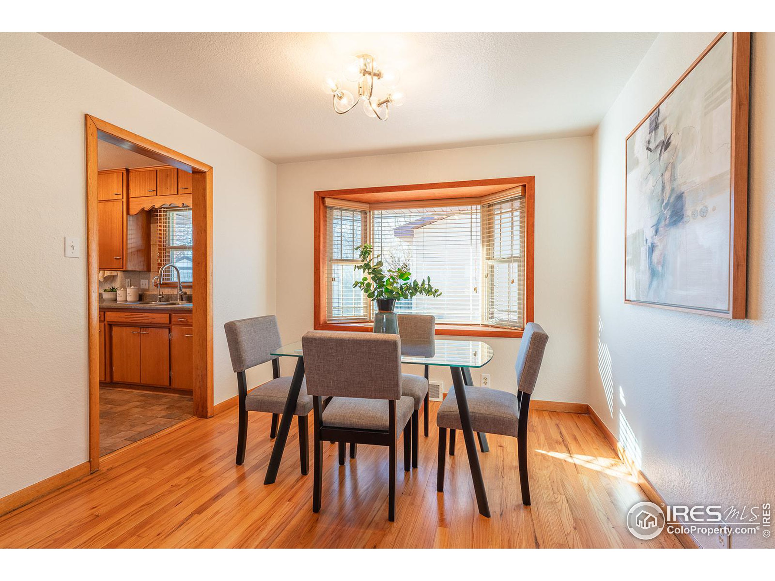 Dining room with a glass table and gray chairs beside a sunlit bay window.