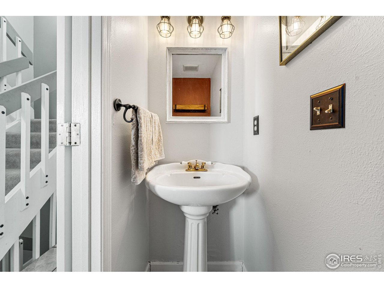 Powder room with a white pedestal sink, brass faucet, and framed mirror beside a carpeted staircase.
