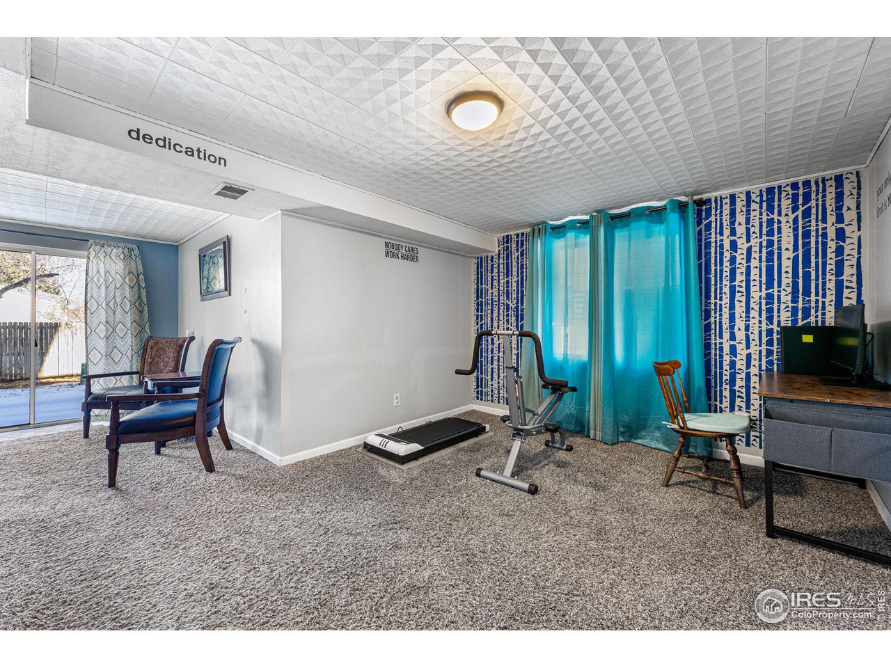 Furnished exercise room with a treadmill, workout machine, turquoise curtains, and patterned wallpaper.