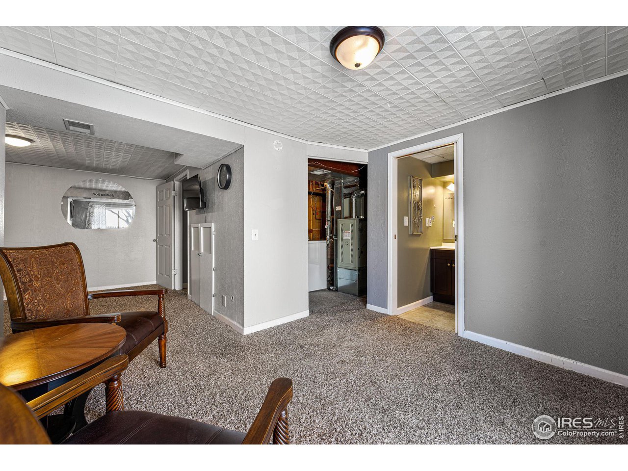 Basement sitting area with patterned armchairs, gray carpet, and open doorways to utility and bathroom areas.